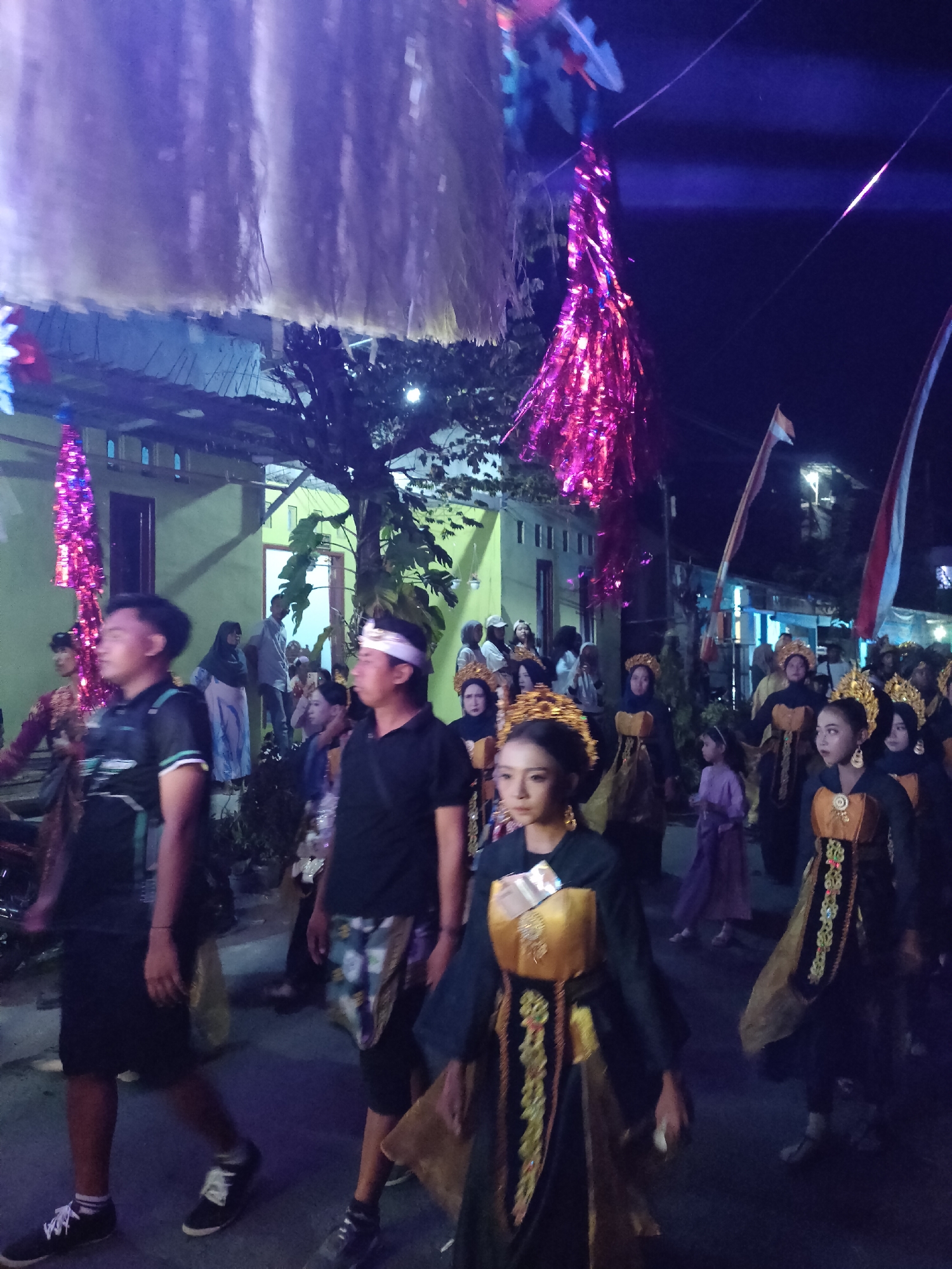 This image depicts a nighttime procession in Tongaswetan, Indonesia, likely a cultural or religious event. The scene is illuminated by decorative lights, with purple and pink streamers hanging overhead, suggesting a festive atmosphere. Several individuals are visible, some participating in the parade while others observe from the side.
In the foreground, two young men are walking. The man on the left wears a black t-shirt with green accents and a backpack, while the man on the right is in a black t-shirt and a patterned sarong. Both are wearing casual footwear.
Further into the procession, a group of young women are dressed in traditional attire. They wear elaborate headdresses and ornate, dark-colored dresses with gold and embroidered accents. Their attire suggests a performance or a significant ceremonial role. Behind them, other participants and onlookers can be seen, including a young girl in a purple dress.
The background reveals a street lined with buildings, some with visible windows and doors. Trees with foliage are interspersed throughout the scene. Two flags, one with red and white stripes and another with white and red colors, are prominently displayed, possibly indicating national or regional affiliation. The overall environment suggests a community gathering in a street setting at night.