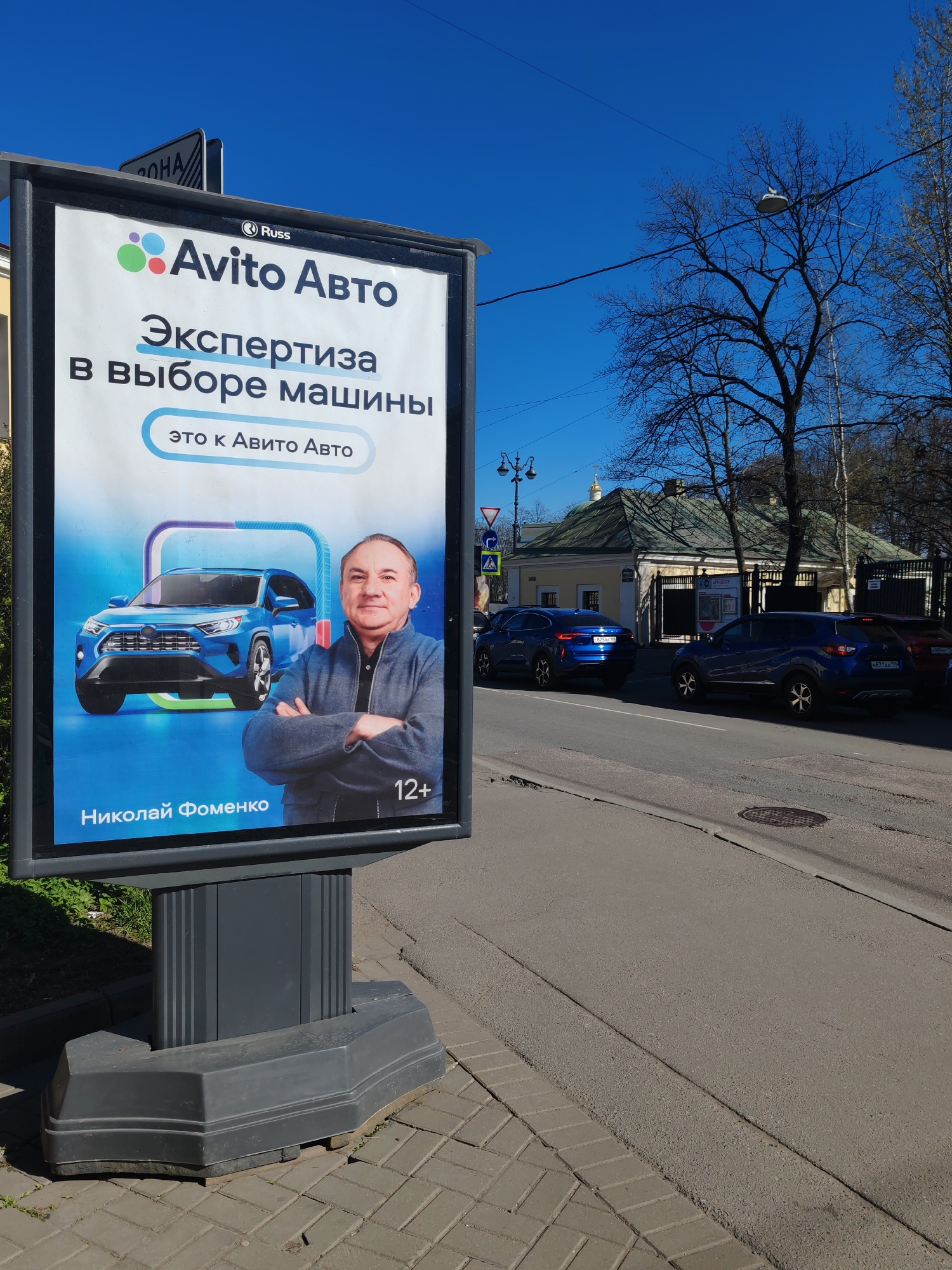The image shows a street scene in what appears to be Saint Petersburg, Russia. A large advertising billboard stands on the sidewalk, promoting "Avito Auto" (a car marketplace). The ad features the slogan "Expertise in car selection - that's Avito Auto," along with an image of a blue SUV and a man identified as Nikolai Fomenko, and a "12+" age rating indication.
In the background, across the street, are several parked cars including blue SUVs, and a yellow building with a green roof can be seen, which is likely a historical building. The sky is clear and blue, suggesting a bright, sunny day.