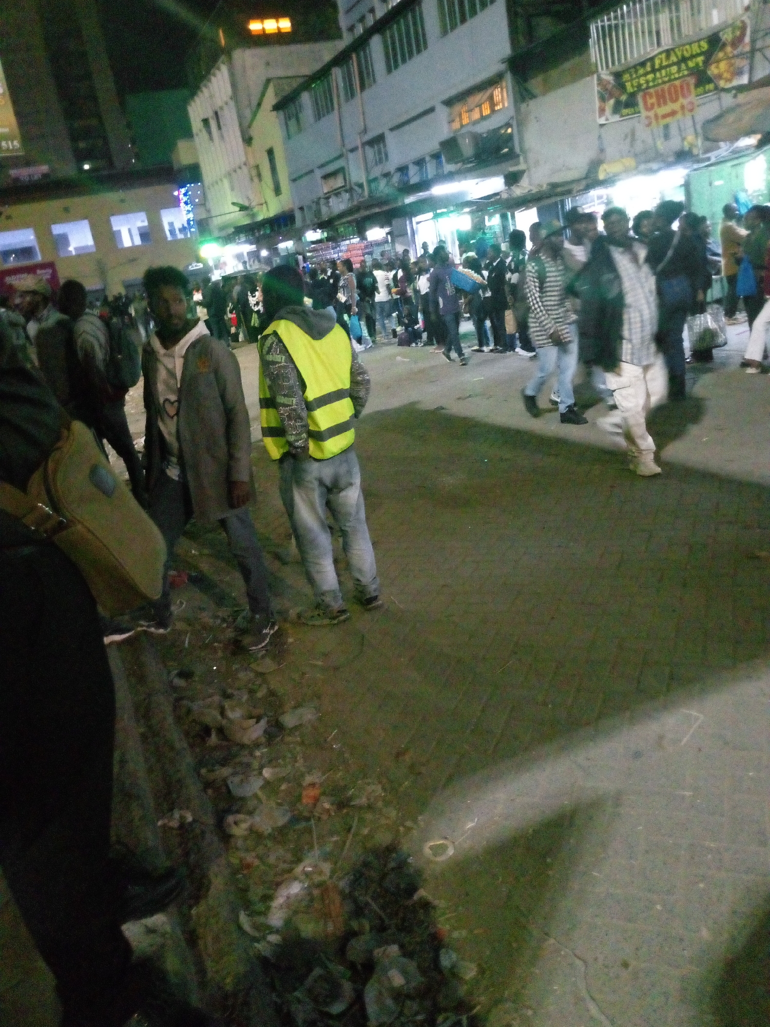 The image depicts a crowded street scene at night in Nairobi, Kenya.
People: A diverse group of individuals is visible. They are mostly dressed in casual clothing, some wearing jackets or sweaters. Several people are walking in the street, while others are standing near buildings. One person is wearing a yellow safety vest.
Environment: The scene is set on a busy street lined with buildings. Streetlights illuminate the area, but overall, the lighting is dim. Some buildings have signs or awnings, one of which reads "FLAVORS RESTAURANT". There is litter and debris visible along the curb and on the street.
Objects: Cars are not visible, indicating the street may be pedestrianized. Backpacks, bags, and general city life items are seen.
Actions: People are walking, standing, and interacting with each other. The general sense is of a bustling urban environment at night.
Location: The street is located in the city of Nairobi, Kenya, as determined by buildings and signage in the photograph.