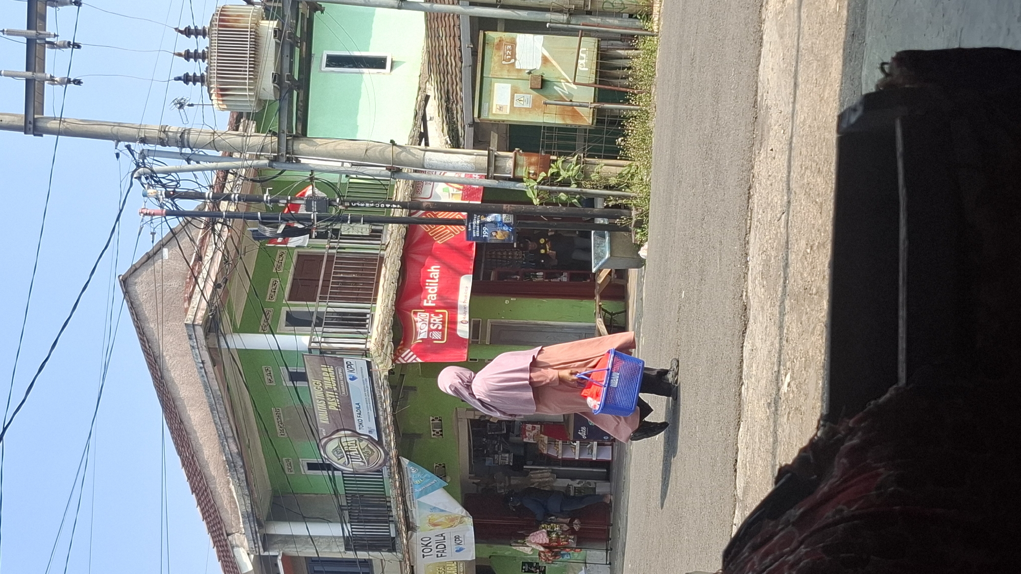 A street scene in Cilaku, Indonesia, shows a woman in a pink hijab and long salmon-colored dress walking to the right across an asphalt road, carrying a blue plastic basket filled with items. She is walking past a row of shops. The shop to her left has a green facade with a sign that reads "TOKO FADILA". To her right, a prominent red banner advertises "SRC FADILAH". Further to the right, there is an electrical transformer on a utility pole. Buildings with green facades and balconies line the street. The sky is clear and blue. In the foreground, a dark object, possibly furniture, partially obscures the lower part of the image.