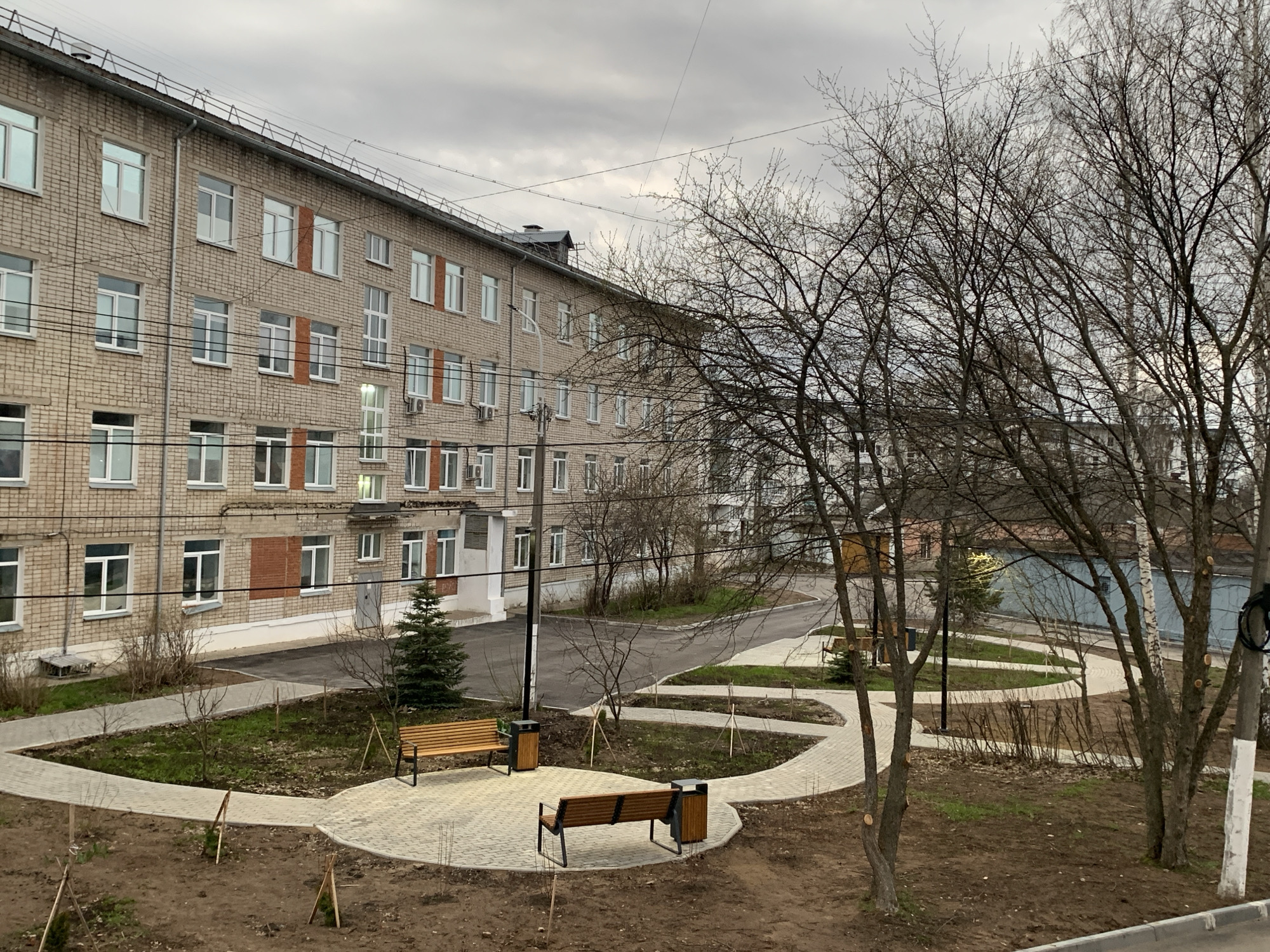 This image depicts an outdoor scene in what appears to be the city of Chaikovsky, Russia. A multi-story brick building with multiple windows and a metal roof is positioned on the left side of the frame. In front of the building, there is a landscaped area with pedestrian pathways constructed from light-colored pavers. Benches and trash receptacles are situated along these pathways. Several young trees, some supported by stakes, are planted throughout the area. The foreground features a view through bare trees with exposed branches, suggesting it is either late autumn or early spring. In the background, there's a road and glimpses of other buildings. The sky is overcast. No people are visible in the image.