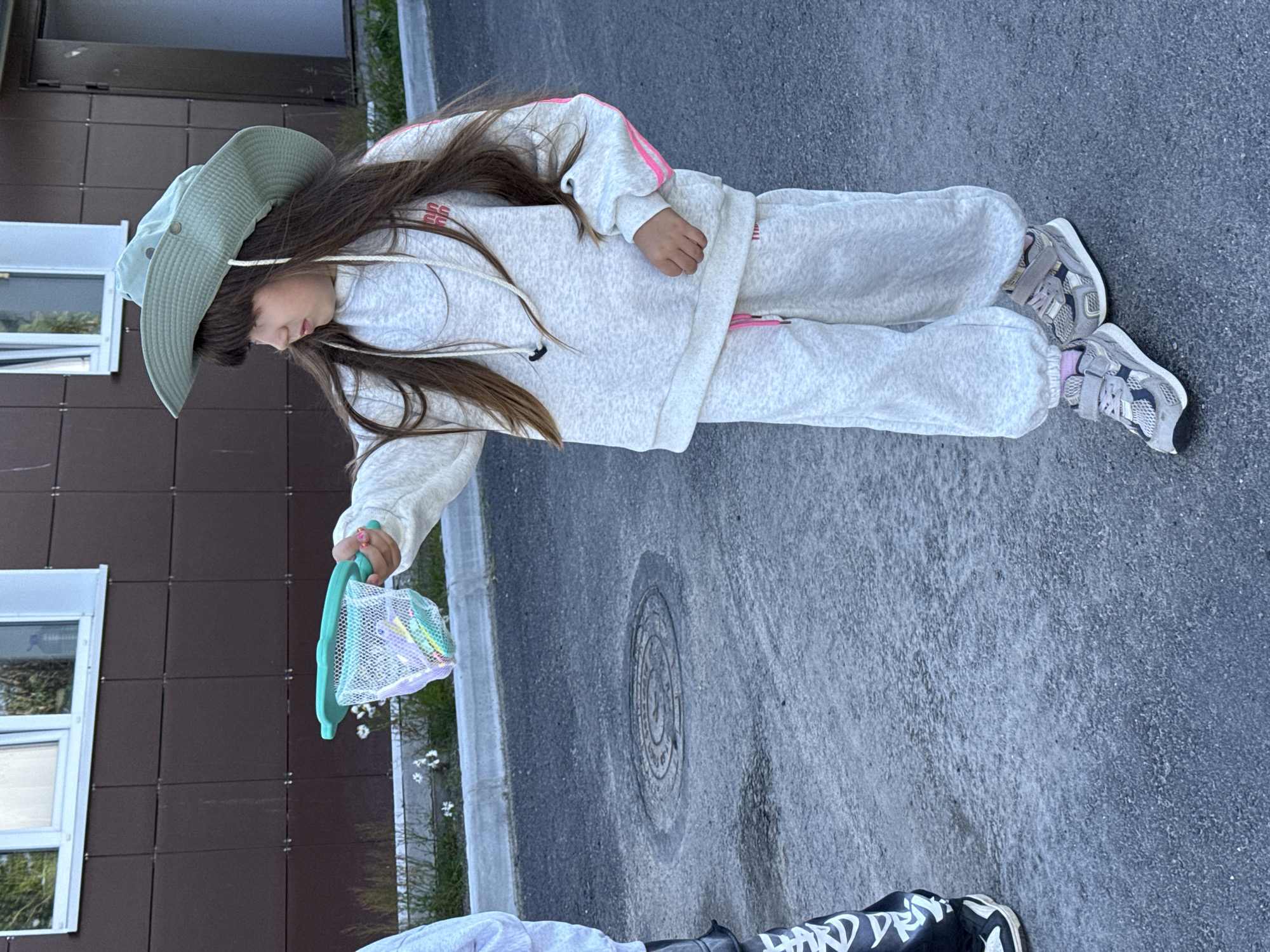 In Teriberka, Russia, a young girl wearing a light grey hooded sweatshirt, matching sweatpants with pink drawstring detail, and a light green wide-brimmed hat, stands on asphalt. She is holding a teal-colored net, catching what appear to be soap bubbles. The net contains small, colorful objects. To the left of the girl, another child's legs are visible, wearing black pants with white graphic lettering and black and white sneakers. Behind the children, a building with dark brown siding and a white-trimmed window is present. The ground is asphalt with a circular manhole cover in the foreground.