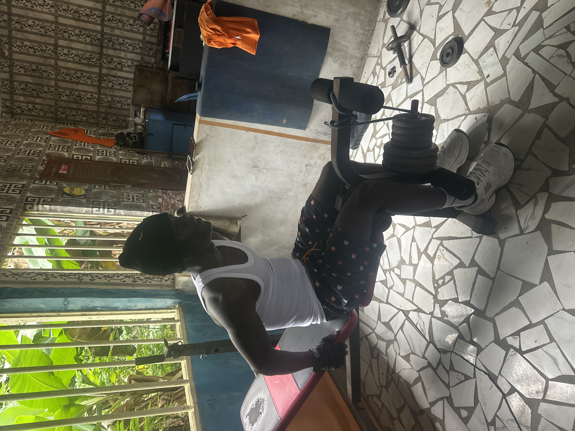 A young Black male wearing a white tank top, black beanie, black shorts with a colorful pattern, and white athletic shoes is performing a leg extension exercise on a black and red weight bench. He is wearing black weightlifting gloves and his feet are strapped into the leg extension apparatus which is loaded with weight plates. The scene appears to be an open-air gym, possibly a home gym, with a tiled floor. In the background, there is a wall decorated with patterned wallpaper, a wooden door, and some blue cabinetry. Large green banana leaves are visible through a barred window to the left of the frame. The gym is located in Badagry, Nigeria.