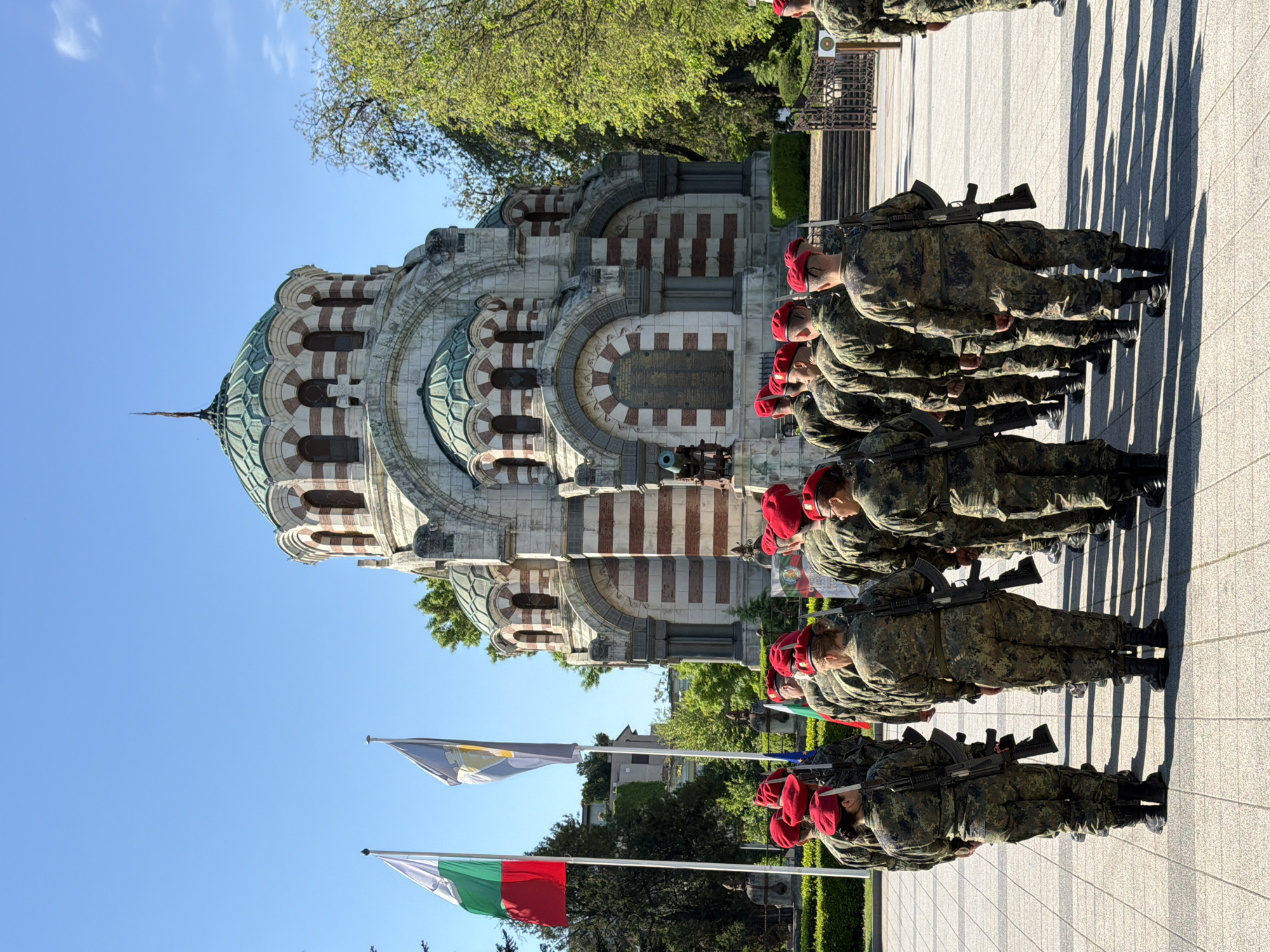 A full shot shows a line of soldiers in camouflage uniforms standing at attention in front of the St. George the Victorious Chapel Mausoleum in Pleven, Bulgaria. The soldiers are wearing red berets and are carrying rifles on their shoulders. The Bulgarian flag and another flag are visible in the foreground. The sky is blue and there are trees in the background. The soldiers are casting long shadows on the pavement.