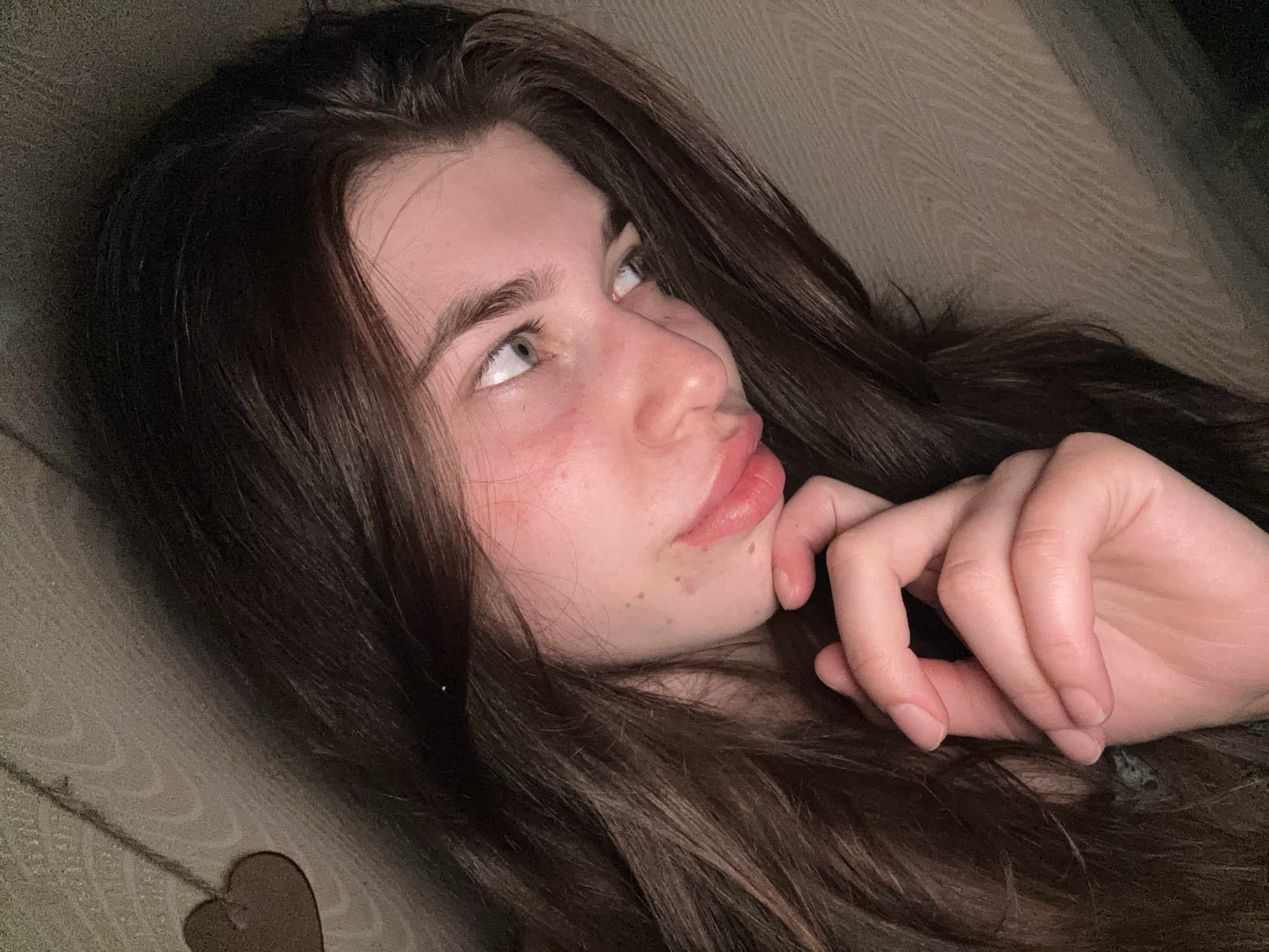 A young woman with long, dark brown hair lies on a beige, textured surface, possibly a sofa or bed. She is facing diagonally towards the upper left corner of the frame. Her left hand is raised near her chin, and her head is tilted back. Her eyes are looking up and to the left. The woman has fair skin with some redness on her cheeks and minor blemishes. A small heart-shaped object is visible at the bottom left of the frame. The location is not discernible.