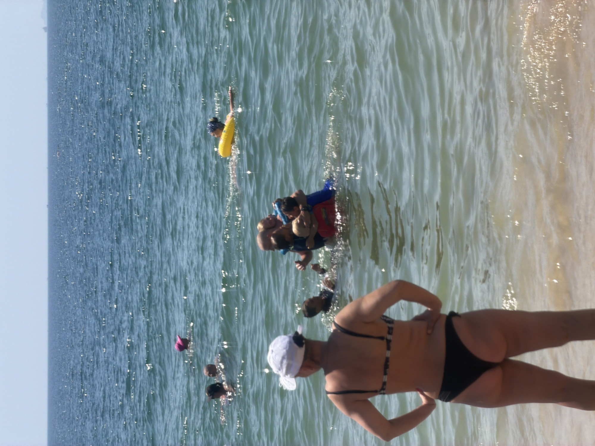 In Nha Trang, Vietnam, on a clear day, several people are enjoying the water at a beach. In the foreground, a woman wearing a white cap and a patterned bikini stands with her back to the camera, facing the ocean. Further out in the water, a group of individuals are gathered. Two children are being held by an adult, and another person is floating on a yellow inflatable ring. In the distance, a few other people are visible swimming or wading in the sea. The ocean is calm with small waves and the sun is reflecting off the water's surface.