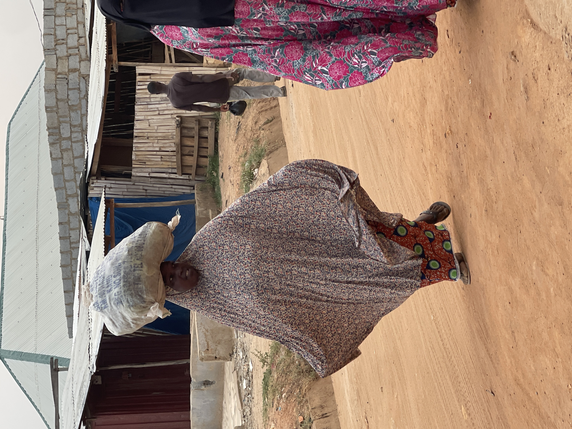 In the Kuchingoro, Nigeria, a woman in a patterned shawl carries a sack on her head, walking on a dirt road. Another woman in a floral patterned dress is visible nearby. A man in dark clothing walks behind them, near wooden buildings. Other buildings with corrugated roofs and blue tarps are also in the background.
