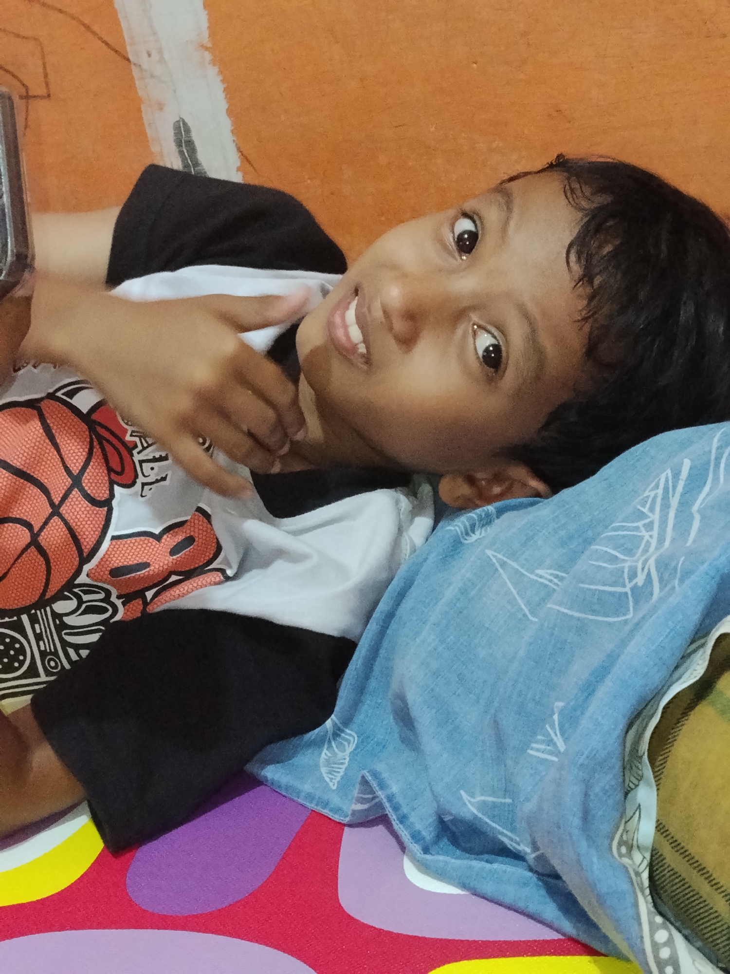 A child is lying on a bed. The child is wearing a black and white shirt with a basketball design. The bed has a colorful patterned sheet and a blue pillow. The wall behind the child is orange. The image was taken indoors, possibly in a home. The specific location in Lhokseumawe, Indonesia, cannot be determined from the image.
