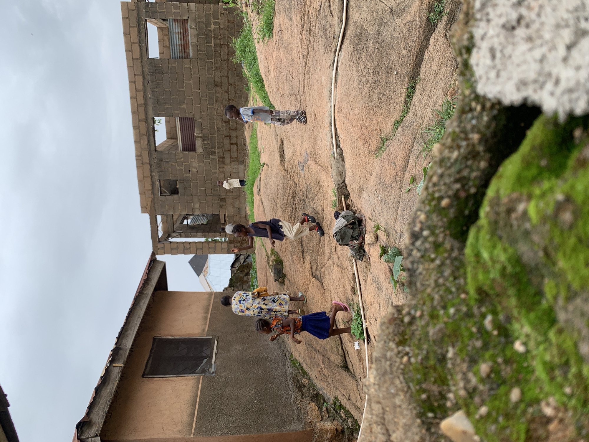 The image depicts an outdoor scene on a rocky, uneven terrain with some greenery. In the foreground, moss-covered rocks are visible. Behind these, a group of children and an adult are present near some buildings. To the left, a partially constructed building with a rough, ochre-colored exterior and a window with a metal grate is seen. Adjacent to it, another building is in a more advanced state of construction, made of cinder blocks with open window frames, some covered with corrugated metal sheets.
Several children are scattered across the rocky ground. One young girl in a blue skirt and an orange and brown top is in motion, appearing to be playing. Another child in a dark blue sleeveless shirt and light-colored pants is also engaged in activity. A third child, wearing a grey t-shirt and patterned pants, stands looking to the right. Further in the background, a person in a white garment stands near the cinder block structure. An adult woman in a patterned dress walks away from the camera, carrying a yellow bag. The sky is overcast. The scene appears to be a residential area undergoing development or in a state of disrepair, possibly in the city of Jos, Nigeria, known for its rocky landscape.