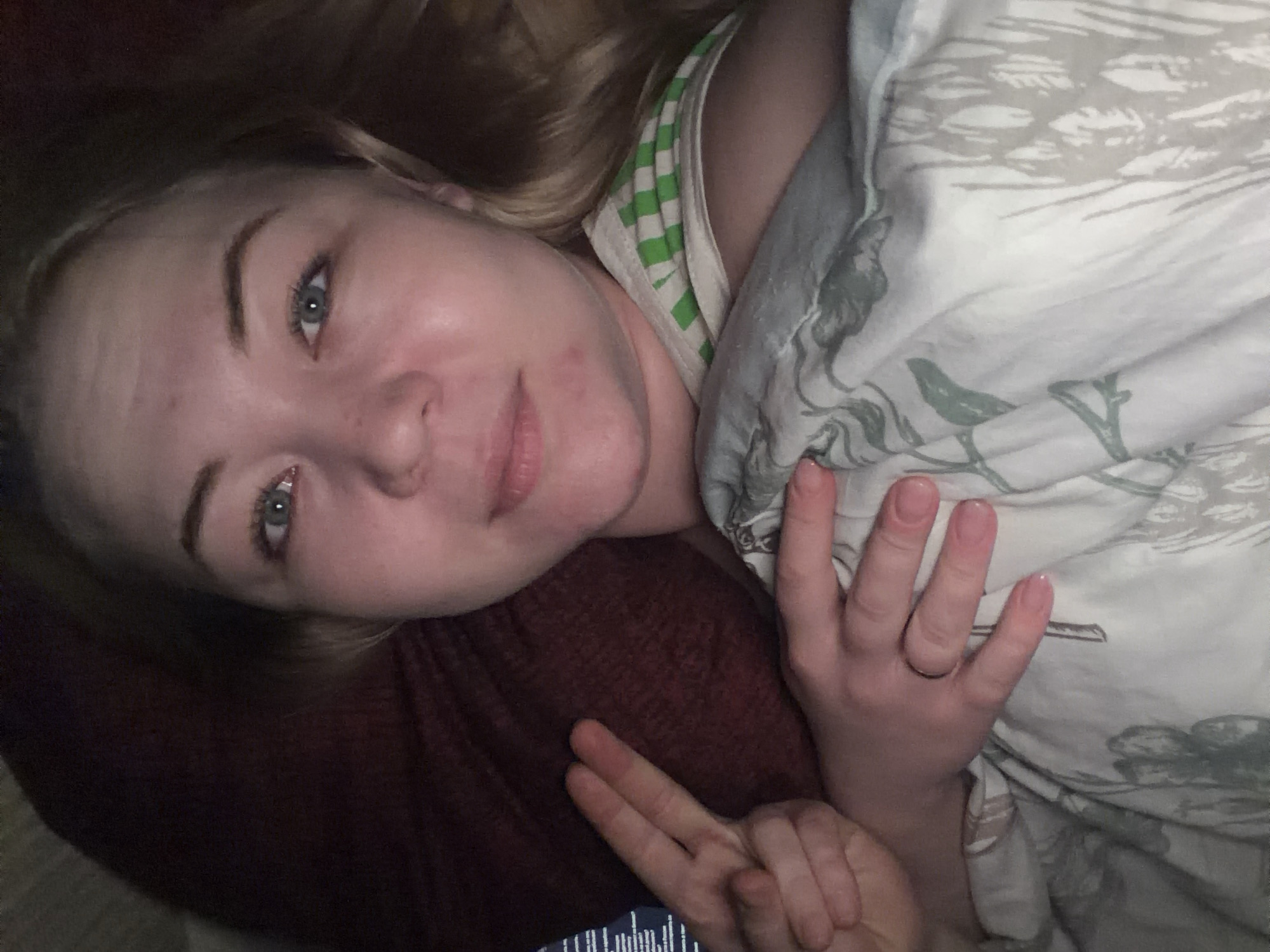 A young woman with blonde hair is lying down, partially covered by a patterned blanket. She is wearing a green and white striped tank top. She is making the peace sign with one hand and holding her other hand up. She has fair skin and blue eyes. The background is dark, suggesting an indoor setting. The location of Noviy, Russia, cannot be determined from this image.