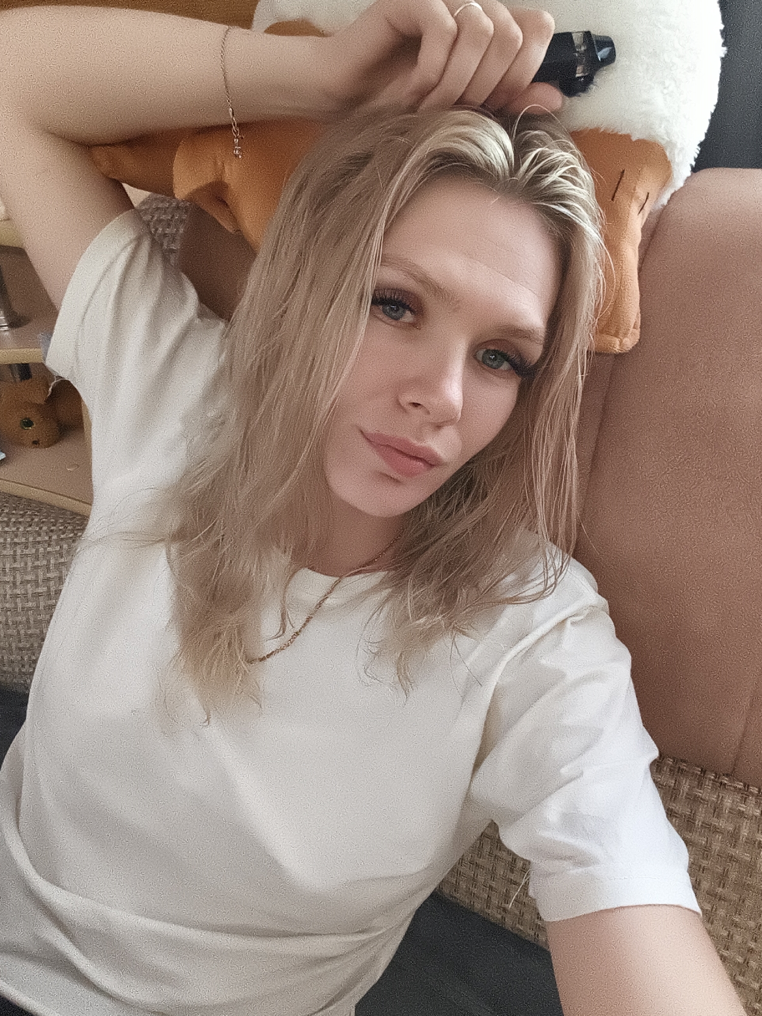 A woman is lying down on a couch in a room. She has long blonde hair and is wearing a white t-shirt and a necklace. Her arms are resting behind her head, and she is looking at the camera. There is a stuffed toy next to her head. The room appears to be a living room with a couch and a woven chair. There is no indication of the specific location within the city of Русский Вожой, Russia.