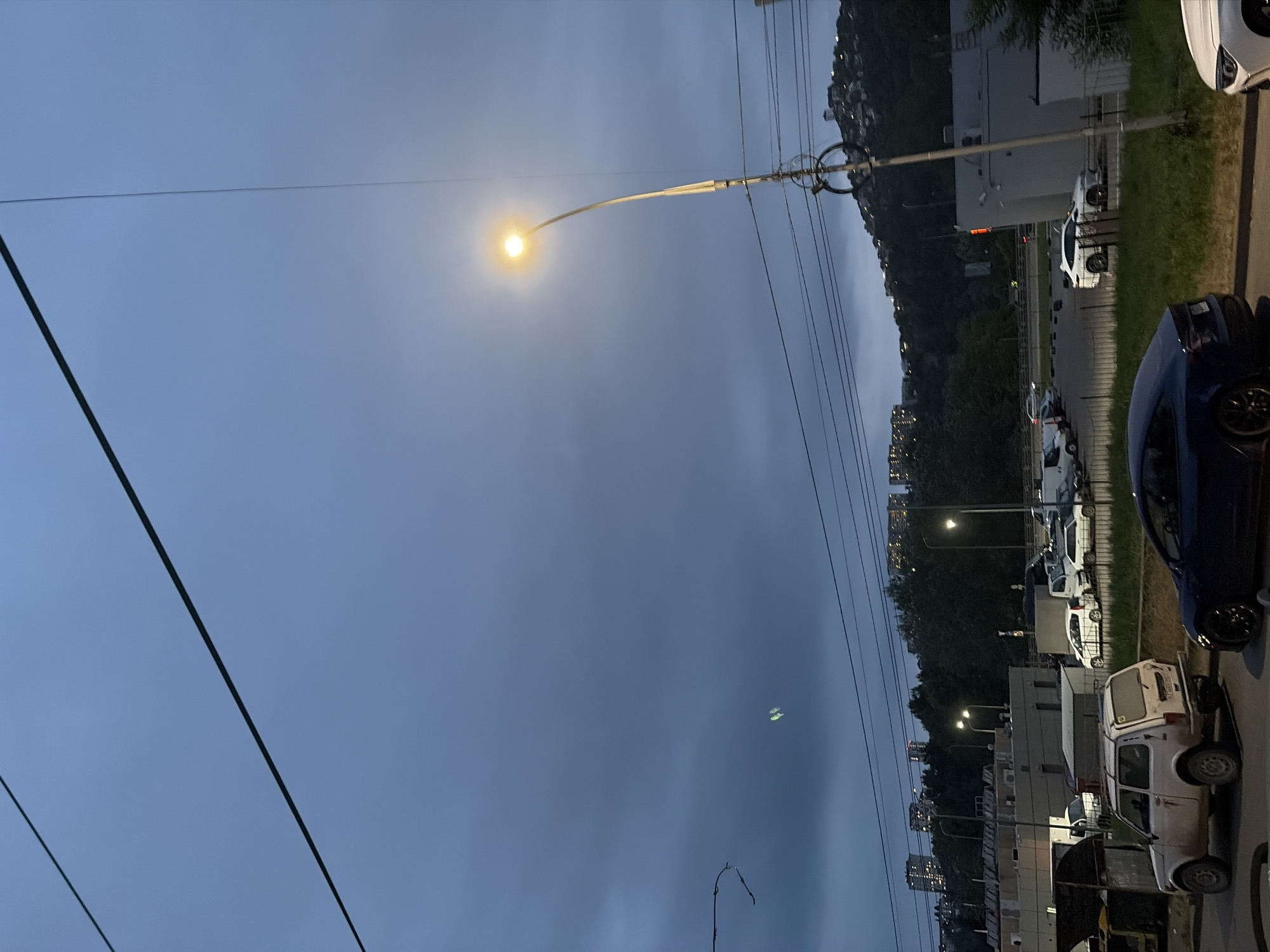 The image depicts an urban street scene in Sochi, Russia, during twilight. The sky is overcast with dark, brooding clouds. A prominent street lamp with a bright yellow light illuminates the foreground, casting a warm glow. Overhead, thick black cables crisscross the sky, obscuring the view of the distant city lights.
In the middle ground, a parking lot is visible with several vehicles, including a white Lada Niva, a dark blue car, and other white passenger cars. Some buildings and trees line the background, with more city lights and structures visible on a distant hillside. The overall atmosphere is somber, with the dim light and heavy clouds creating a moody ambiance.