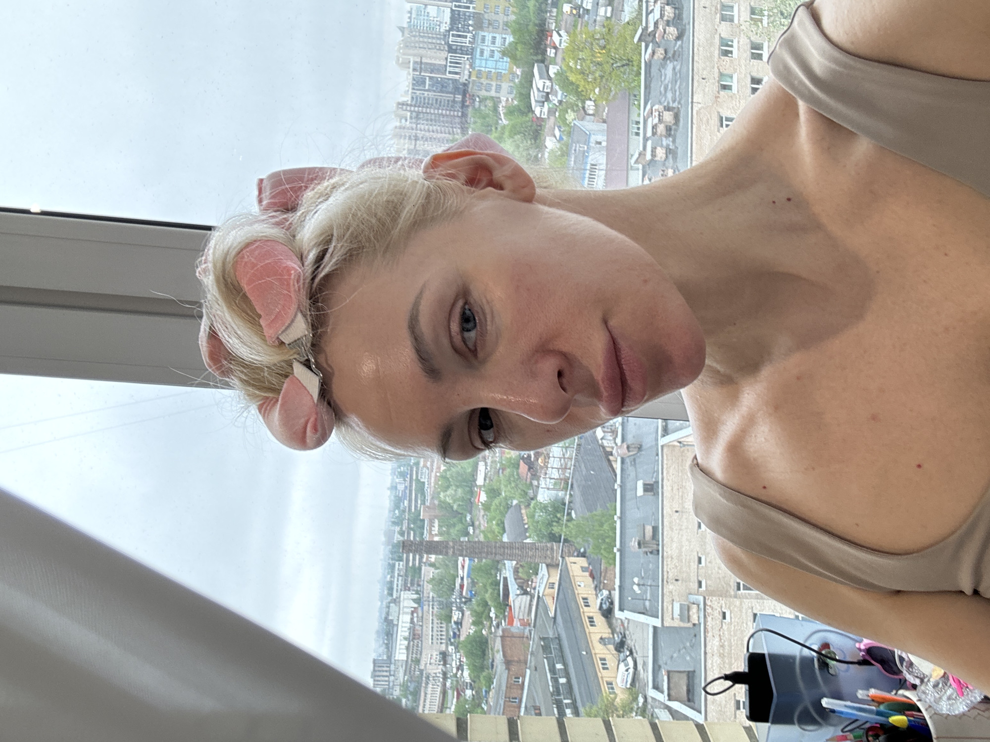 A woman with pink hair rollers is looking toward the camera. She is wearing a beige tank top. In the background, there is a city with high-rise buildings and some greenery. A desk with pens and other items is also visible. The woman appears to be indoors, possibly in an apartment with a view of the city.