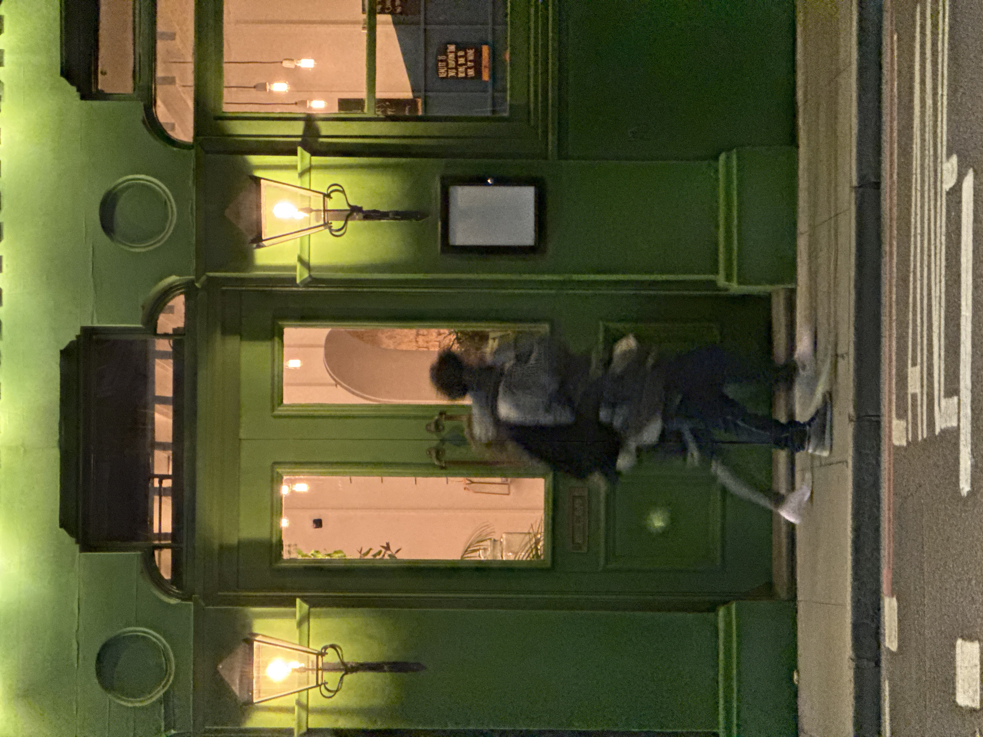 A nighttime photograph depicts the exterior of a green building in London, United Kingdom. Two people are blurred in motion in front of the building. The building features architectural details such as arched windows, decorative sconces with illuminated lights, and a rectangular sign. A traffic sign is visible on the street.