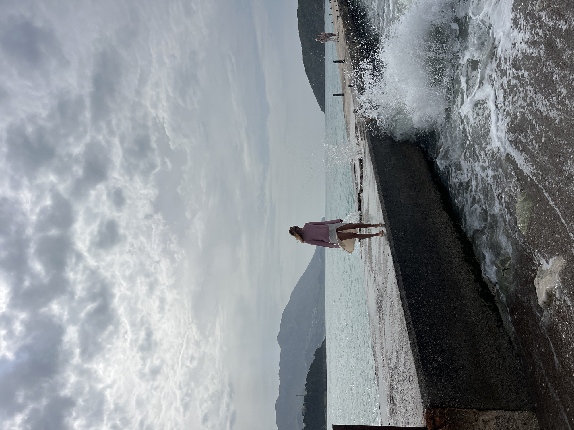 A woman stands on a pier in Budva, Montenegro. She faces the sea with her back to the mountains in the background. She is wearing a light-colored skirt and a pink sweater. Waves are crashing on the pier. The sky is cloudy.