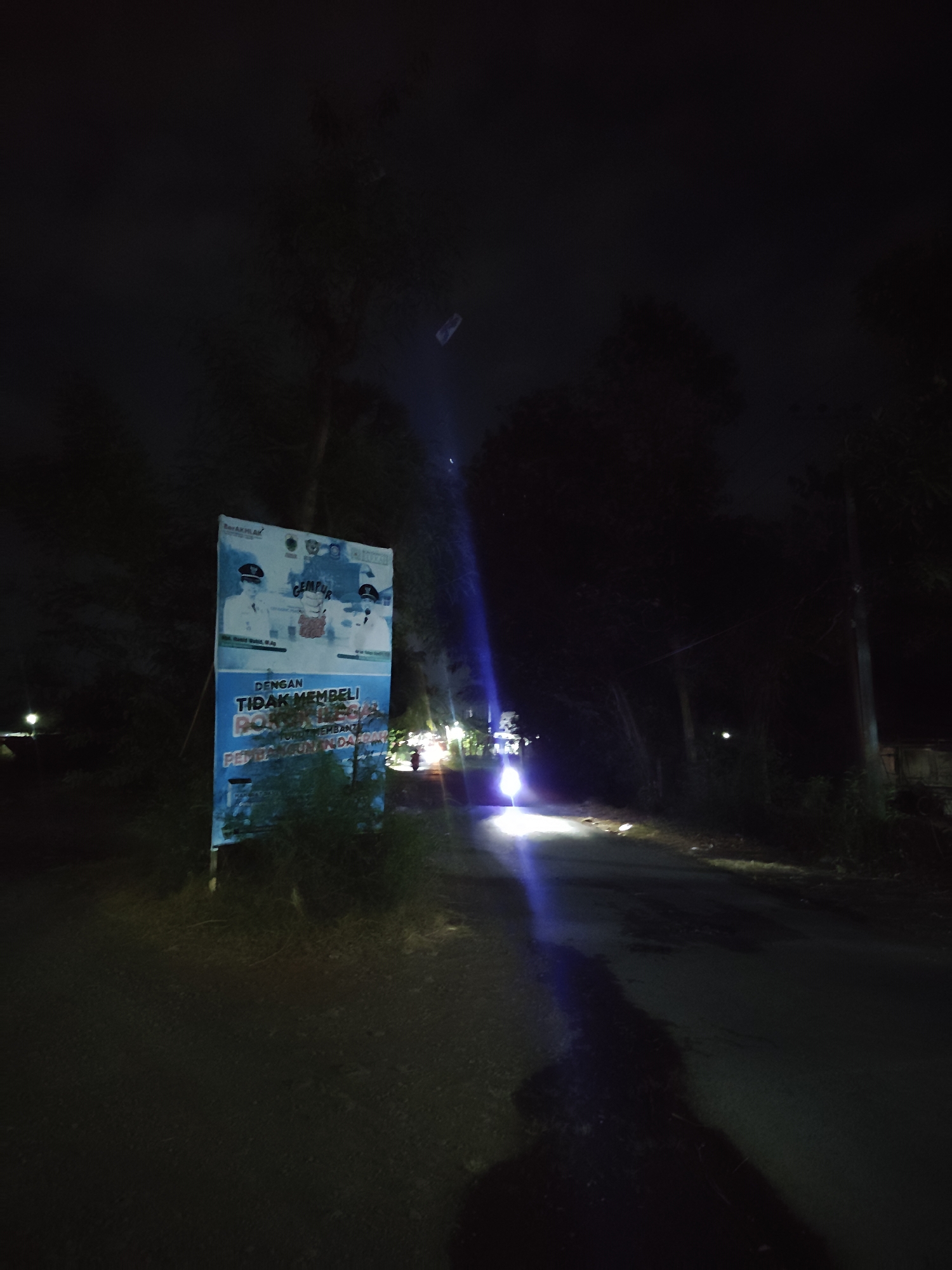 The image depicts a nighttime scene on a rural road in Suling Wetan, Indonesia. A prominent blue and white banner stands on the left side of the road, displaying official portraits and text in Indonesian. The banner appears to be a public service announcement or advertisement. In the center of the image, a motorcycle with its headlights on is approaching, casting a bright beam of light onto the road and creating a strong lens flare. The surrounding environment consists of dark trees and foliage, with a few distant lights suggesting other structures or activity. The road surface is asphalt and appears wet in some areas. The overall atmosphere is dark and somewhat mysterious due to the low light conditions and the presence of artificial light sources.