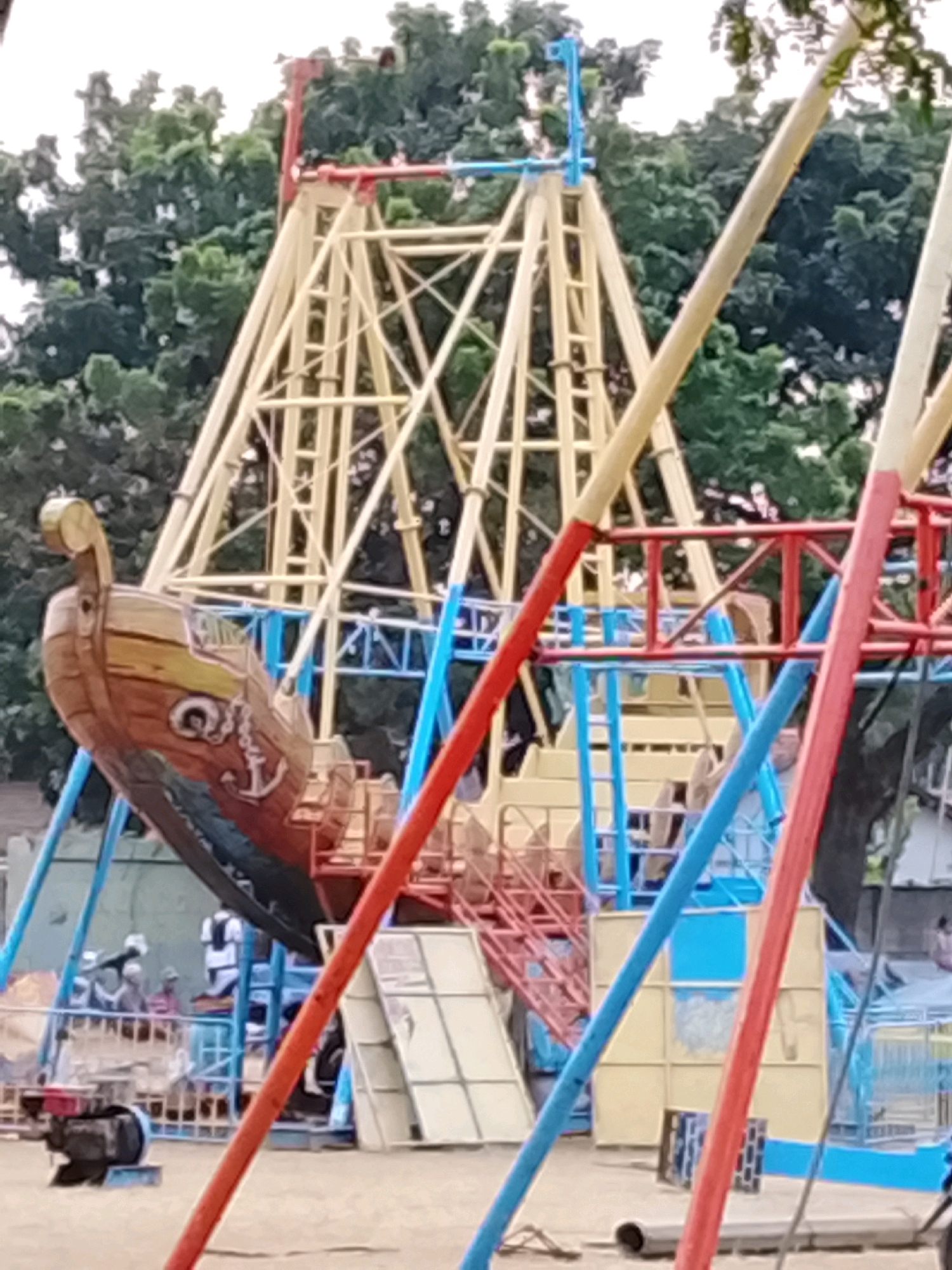 The image depicts a pirate ship amusement ride at a park. The ship is brown with yellow accents and has an anchor and eye painted on its side. It is suspended from a yellow metal frame, which is partially obscured by red and blue poles supporting the ride. In the background, trees with green foliage are visible. Several people are present in the park, with some appearing to be waiting to ride the attraction. The ground is sandy. The scene suggests a public recreational area, possibly an amusement park or a fairground in Harjosari Lor, Indonesia.