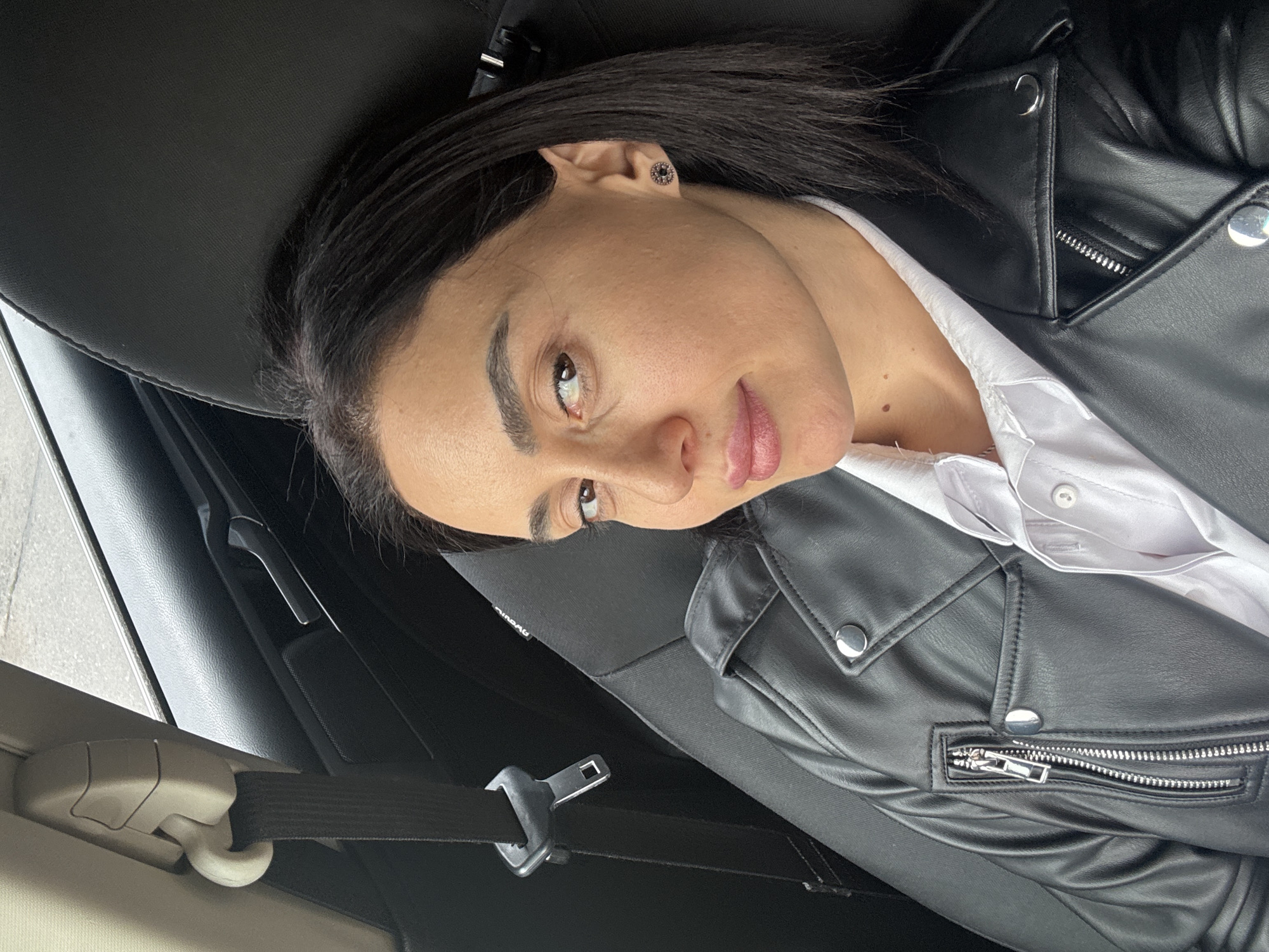 A woman is lying in the back seat of a car. She is wearing a black leather jacket over a white shirt. She has dark hair and is wearing earrings. The seatbelt is fastened across her. The background suggests a car interior. The location cannot be determined from the image.