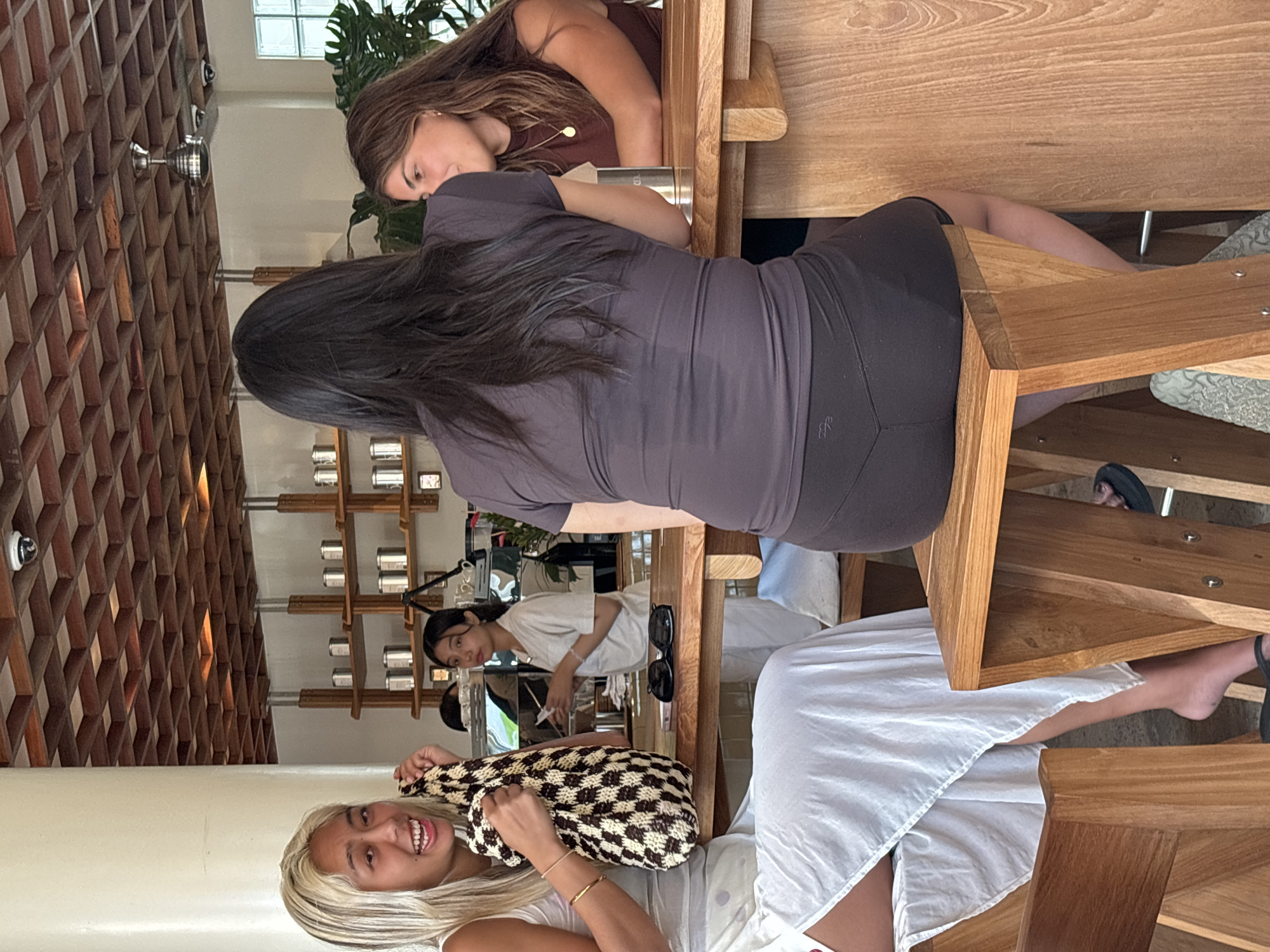 Here is a detailed description of the image:
The image appears to be taken at a cafe or restaurant in North Kuta, Indonesia. The interior features a textured wooden wall and a display of various jars or containers.
Several individuals are present in the scene. Three women are seated at a wooden table. One has blonde hair and is smiling, holding a patterned bag. Another woman, seated beside her, is wearing a white dress or robe. The third woman, seated closer to the top of the image, has dark hair and wears a purple top and dark leggings. A fourth woman is visible in the background, near the display shelves, wearing a white shirt.
The scene is well-lit, suggesting it is daytime. The focus is on the people at the table and the overall ambiance of the cafe.