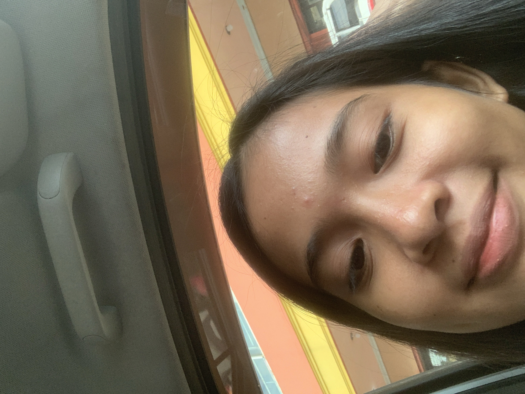 The image depicts a young woman with dark hair, likely in her late teens or early twenties, taking a selfie from inside a vehicle. The vehicle's interior is visible, with a gray headliner and a grab handle. Through the car window, there are exterior building structures and colors visible, including yellow, orange, and white elements, suggesting a built environment of shops or homes. The lack of identifying landmarks makes it impossible to confirm the location within Samarinda, Indonesia. The woman's expression is a slight smile, and she is looking at the camera. The scene appears to be in daylight.