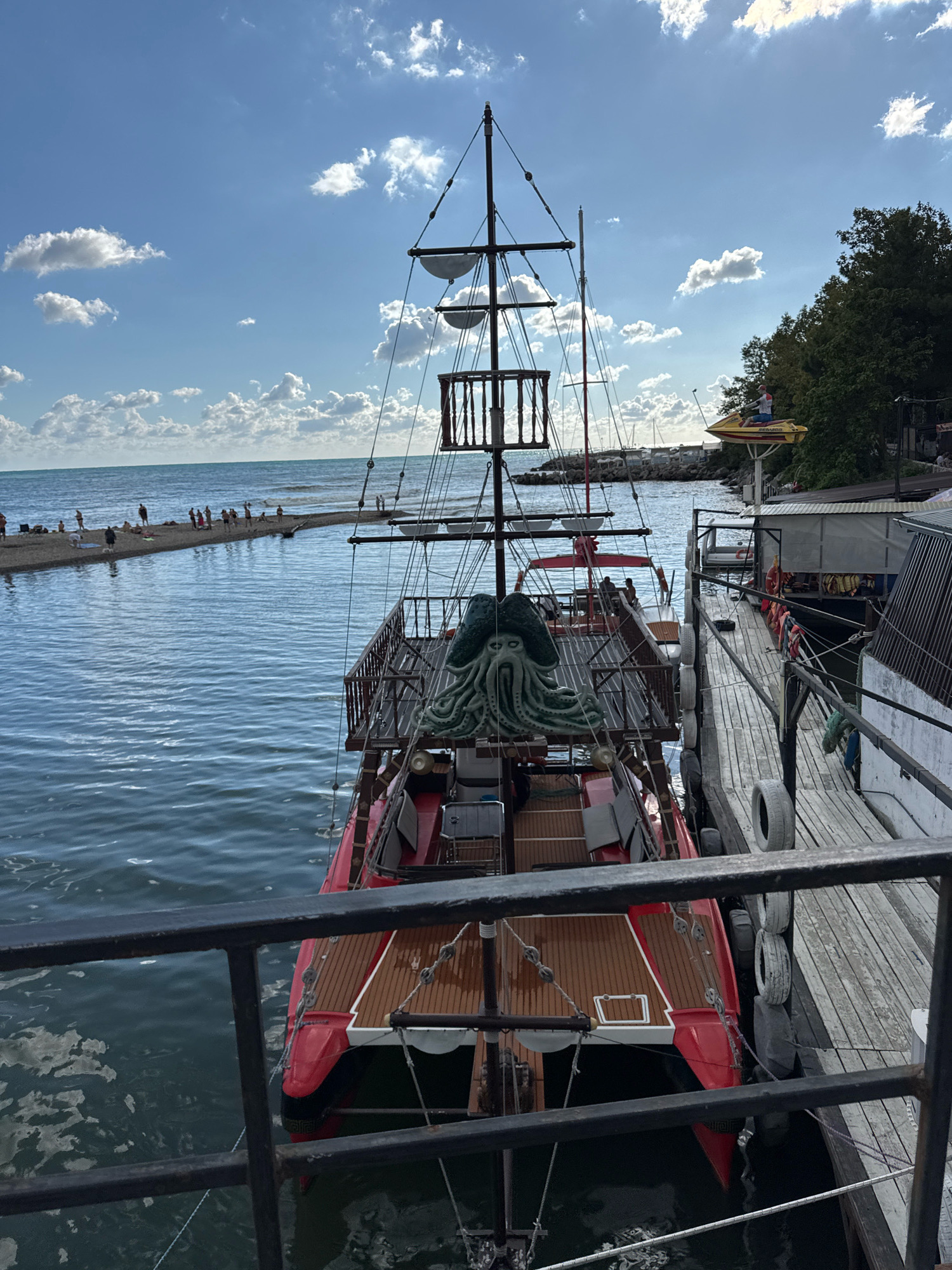 The image depicts a sunny day at a seaside location in Novomikhaylovsky, Russia. In the foreground, a black metal railing overlooks a body of water. To the right of the railing is a wooden pier with several tires acting as fenders. Moored next to the pier is a red catamaran-style boat with a wooden deck, resembling a pirate ship. A green octopus sculpture is visible on the boat's deck. In the middle ground, the sea extends towards a sandy spit where several people are sunbathing and wading. The horizon shows a calm sea meeting a bright blue sky with scattered white clouds. On the right side of the image, behind the pier, there are buildings with awnings and a water slide structure with a figure on a yellow jet ski. Trees line the background on the far right.