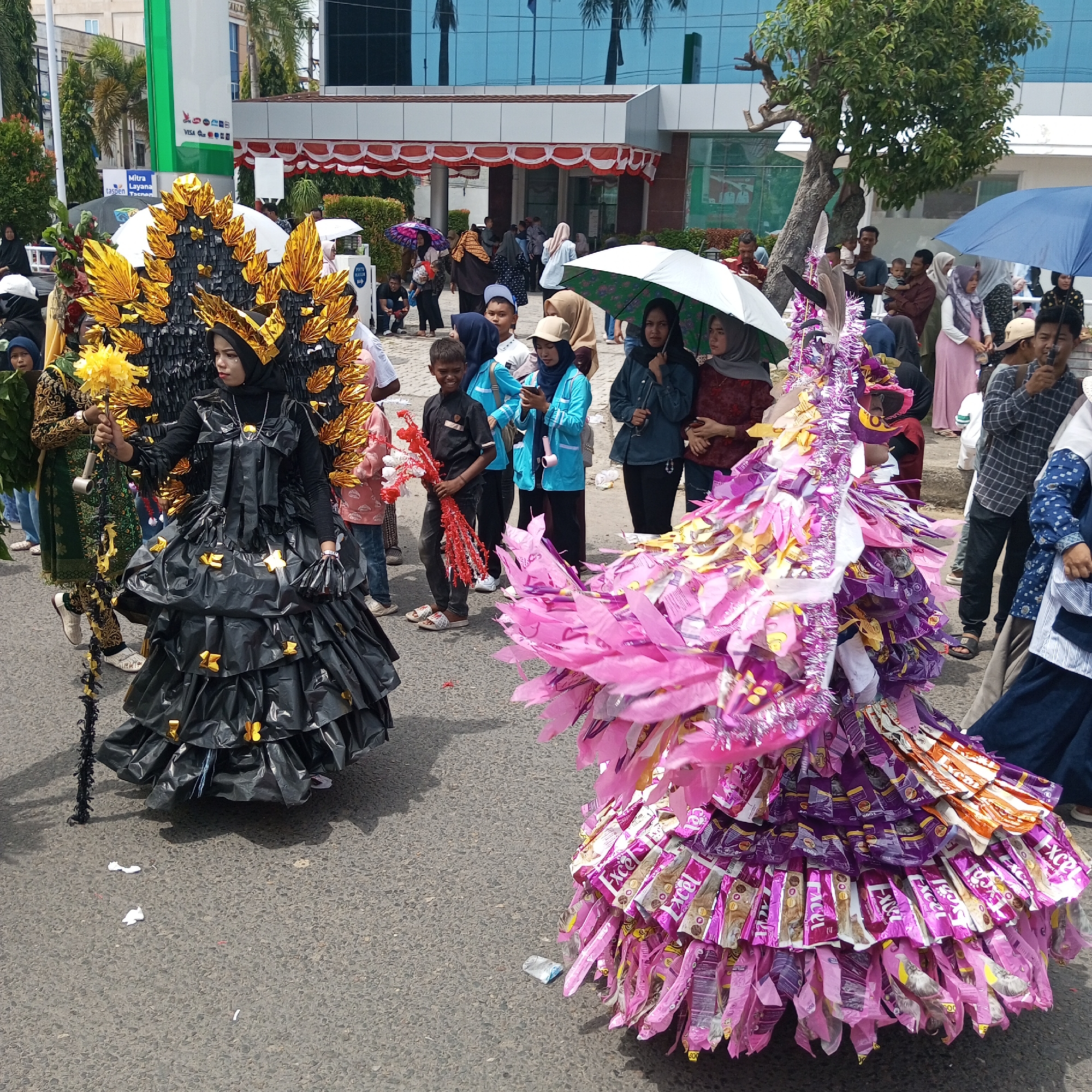 The image depicts a street scene in Lhokseumawe, Indonesia, featuring a parade or cultural event. Two individuals are prominently featured in elaborate costumes. The person on the left wears a black, layered, ruffled costume with golden, fan-like headwear and a golden flower scepter. They are dressed in a black headscarf and black attire beneath the costume. To the right, another individual is adorned in a voluminous pink and purple costume constructed from what appear to be repurposed packaging, possibly candy wrappers, with dangling strips of material. Both individuals are walking on a paved street, which is lined with spectators of various ages. Some spectators are holding umbrellas, suggesting it may be sunny or light rain. In the background, a modern building with blue glass windows and a white facade stands, along with trees and street furniture. The overall atmosphere appears festive and public.