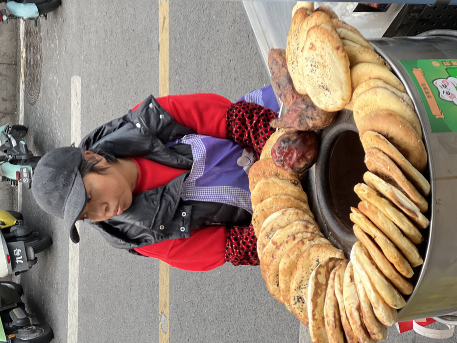 This image depicts a street vendor operating a food stall on an asphalt road in Yuzhong District, China. A single individual, an adult female, is positioned behind a round, stainless steel food cart in the foreground. She wears a black baseball cap, a black sleeveless puffer vest over a red long-sleeved shirt, and a purple and white checkered apron. Red and black patterned sleeves are visible at her wrists. Her gaze is directed towards the left, and her expression is neutral with lips slightly pursed.
The food cart displays a circular arrangement of numerous baked flatbreads, predominantly golden-brown, some featuring dark seeds (possibly sesame) on their surface. These flatbreads are stacked vertically around a central circular opening in the cart, which is empty. Interspersed among the flatbreads are three dark, irregularly shaped roasted tubers, consistent with sweet potatoes or yams. A green sticker with a white cartoon cat and Chinese characters is affixed to the lower right side of the cart.
The background shows an asphalt street with a faded yellow painted line. Several electric scooters or small motorcycles are parked along the curb. One scooter has a white license plate displaying the Chinese characters "九号" (Number Nine). Another light blue scooter and a yellow scooter are partially visible. A circular manhole cover is also discernible on the road surface. The overall lighting suggests an overcast or diffused daytime condition.