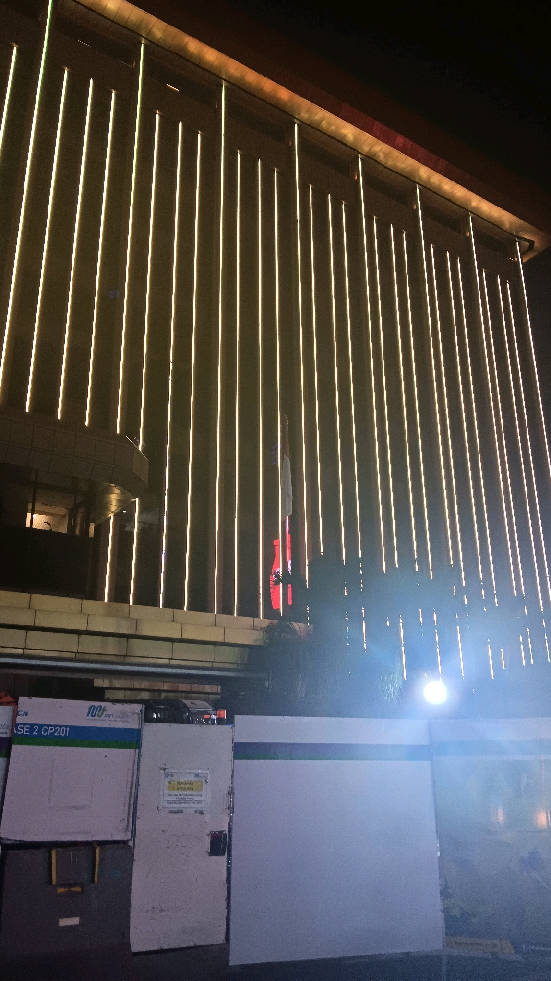 The image shows the exterior of a building at night, illuminated by vertical lines of white lights. In the lower part of the image, there are several metal and white panel structures, possibly part of a street-level entrance or a construction site. One of the panels displays signage, including "CN SE 2 CP201" and a logo with "MRT Jakarta". Another panel has a yellow sign with Indonesian text. The building's facade is modern, with a sleek design emphasized by the lighting. The overall scene appears to be in an urban setting. The image location is the MRT Jakarta station in Central Jakarta, Indonesia.