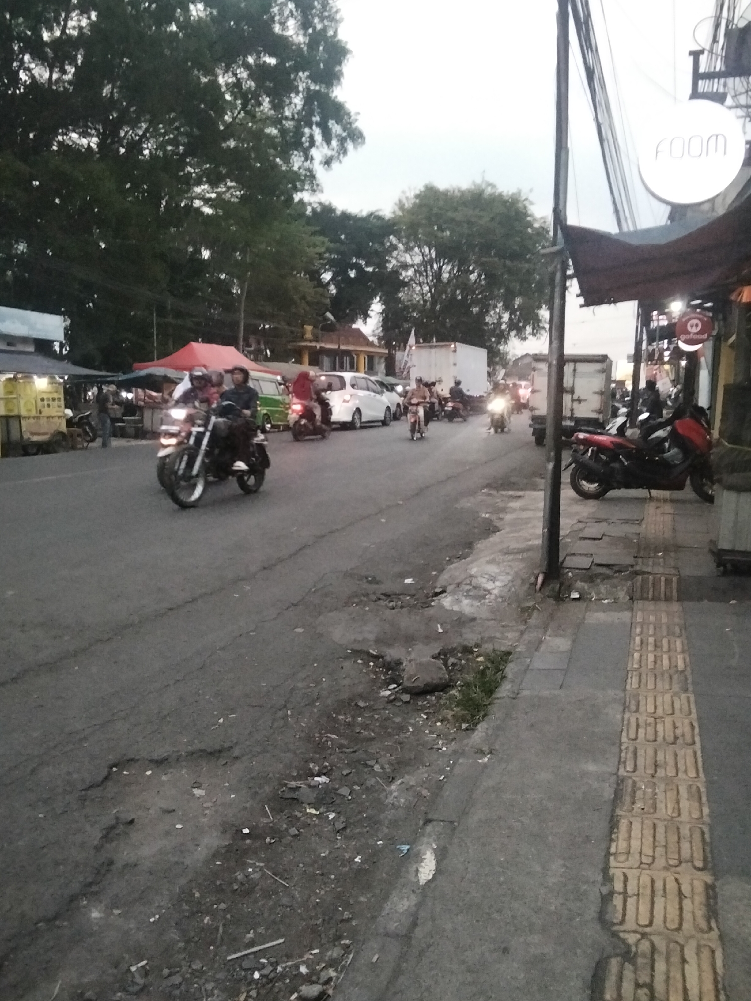 The image depicts a street scene in Sukamanah, Indonesia, likely during dusk or early morning due to the subdued lighting. The foreground is dominated by a paved road with visible cracks and debris. A sidewalk runs along the right side of the road, featuring tactile paving for visually impaired pedestrians. Several motorcycles are present on the road; one prominently features two riders wearing helmets, its headlights illuminated. Other motorcycles and a few cars, including a white SUV and a green van, are also visible further down the street. On the left side of the road, stalls or shops are set up, one with a red tarp overhead. A large tree with dense foliage is on the left, and a light pole stands near the sidewalk. A white circular sign with the text "foom" is mounted on a pole above the shops. The sky is overcast, contributing to the diffused light.