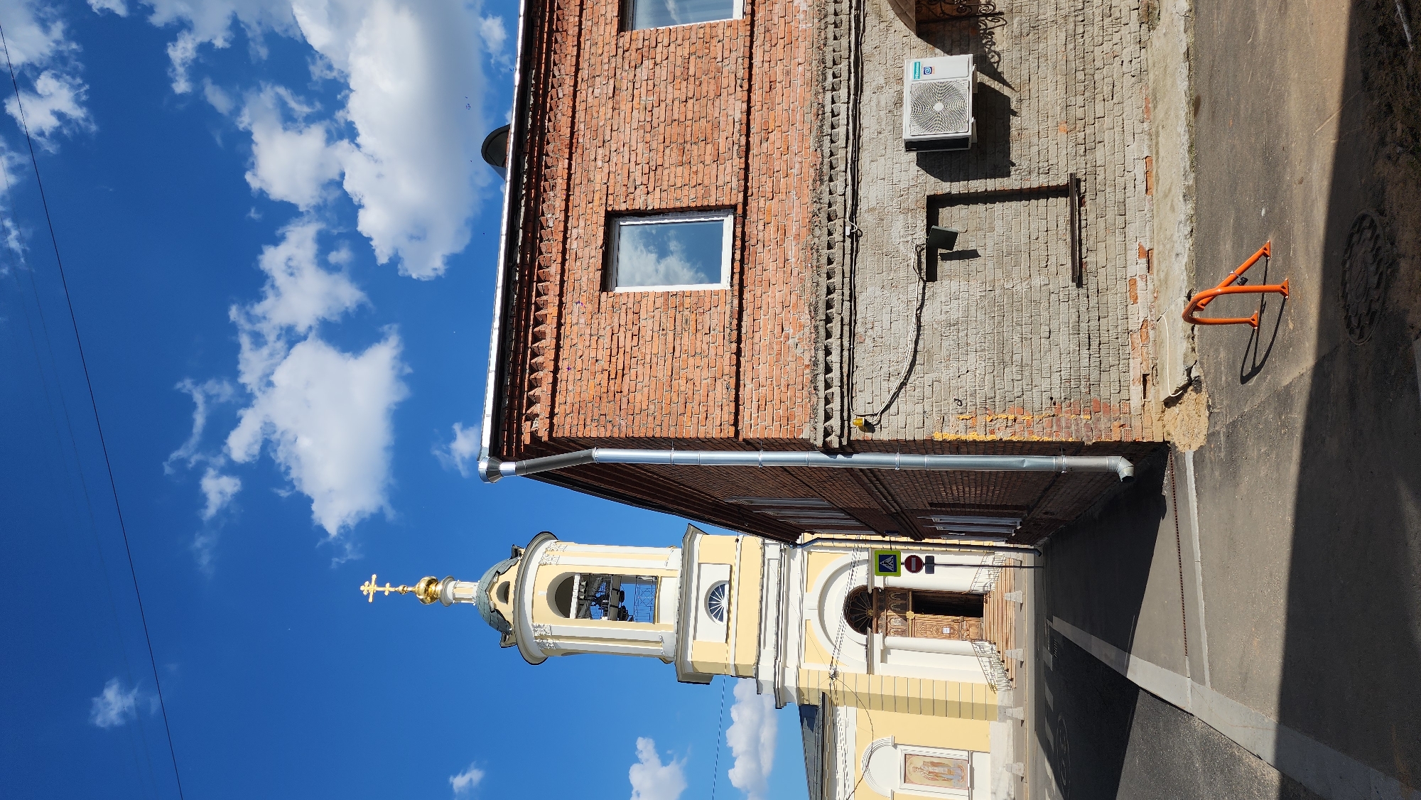 The image depicts a sunlit urban street scene, characteristic of an old Russian city like Moscow, on a clear day with a bright blue sky and scattered white cumulus clouds.
The primary subject is a light yellow church or chapel, positioned on the left side of the frame. It features a prominent bell tower with arched openings, crowned by a golden dome and a golden Orthodox cross. The church's entrance, visible beneath the tower, has decorative wooden doors and is flanked by columns. Near the entrance, two road signs are visible: a triangular pedestrian crossing sign and a red circular sign.
To the right, a two-story brick building stands tall, displaying textured reddish-brown and older grey brickwork. It has at least two windows with white frames, reflecting the sky and clouds. An air conditioning unit is mounted on the upper part of the building, and a metallic downspout runs vertically along its facade.
The foreground shows an asphalt street or alleyway. Strong, well-defined shadows are cast by the buildings, the air conditioner, and an orange metal structure on the pavement, indicating bright, direct sunlight, likely midday or early afternoon. The orange metal structure is possibly a bicycle rack or a barrier. A circular manhole cover is also visible on the street surface.
The scene is devoid of people or any explicit activity, conveying a calm and peaceful urban moment.