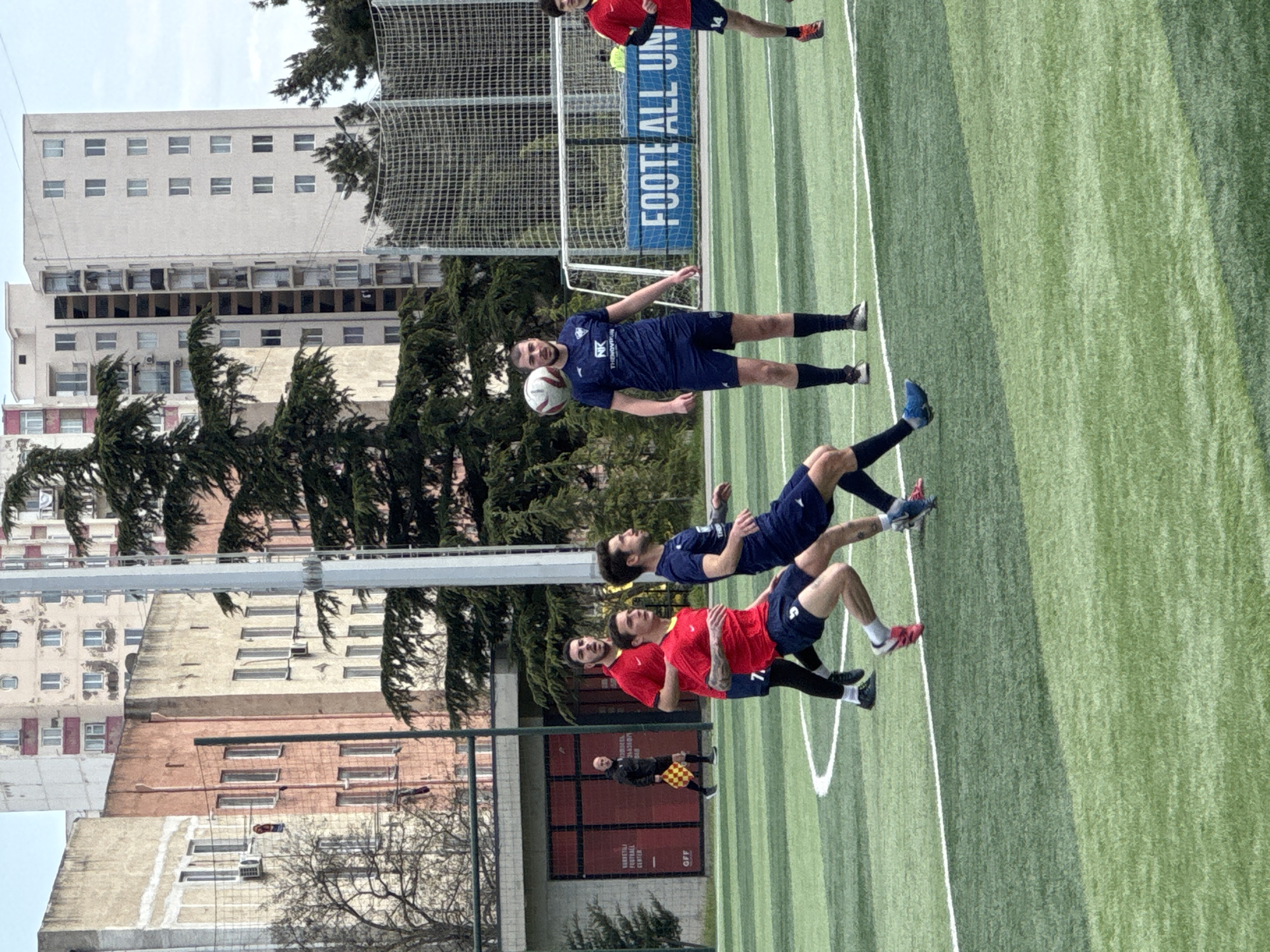 This image shows a soccer game in progress on a green artificial turf field. Four players are visible: three wearing red shirts and one wearing a dark blue shirt. The blue-shirted player is heading a soccer ball, while the other players are in motion. In the background, a building and a net with "FOOTBALL UNITED" printed on it are visible. An official is also present in the background, holding a checkered flag. Buildings characteristic of Tbilisi, Georgia are visible in the background.
