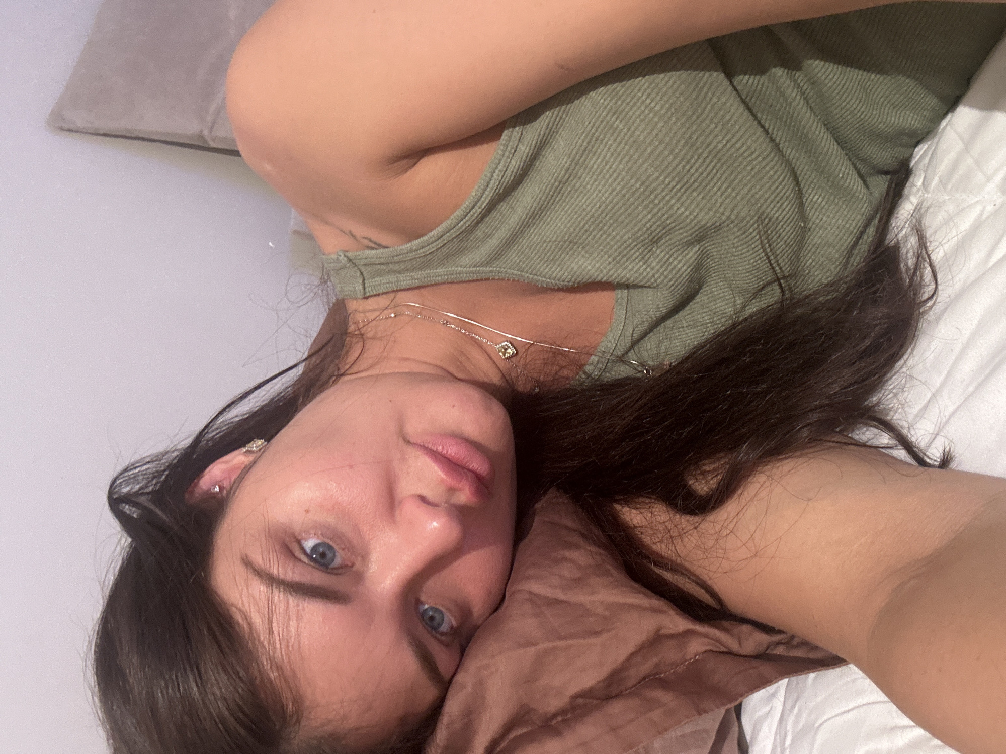 A woman is lying on a bed, resting her head on a brown pillow. She has fair skin, dark brown hair, and blue eyes. She is wearing a light green tank top and a silver necklace with a pendant. A grey pillow is positioned behind her upper back. The bedsheets are white and quilted. The photo's location is not discernable.