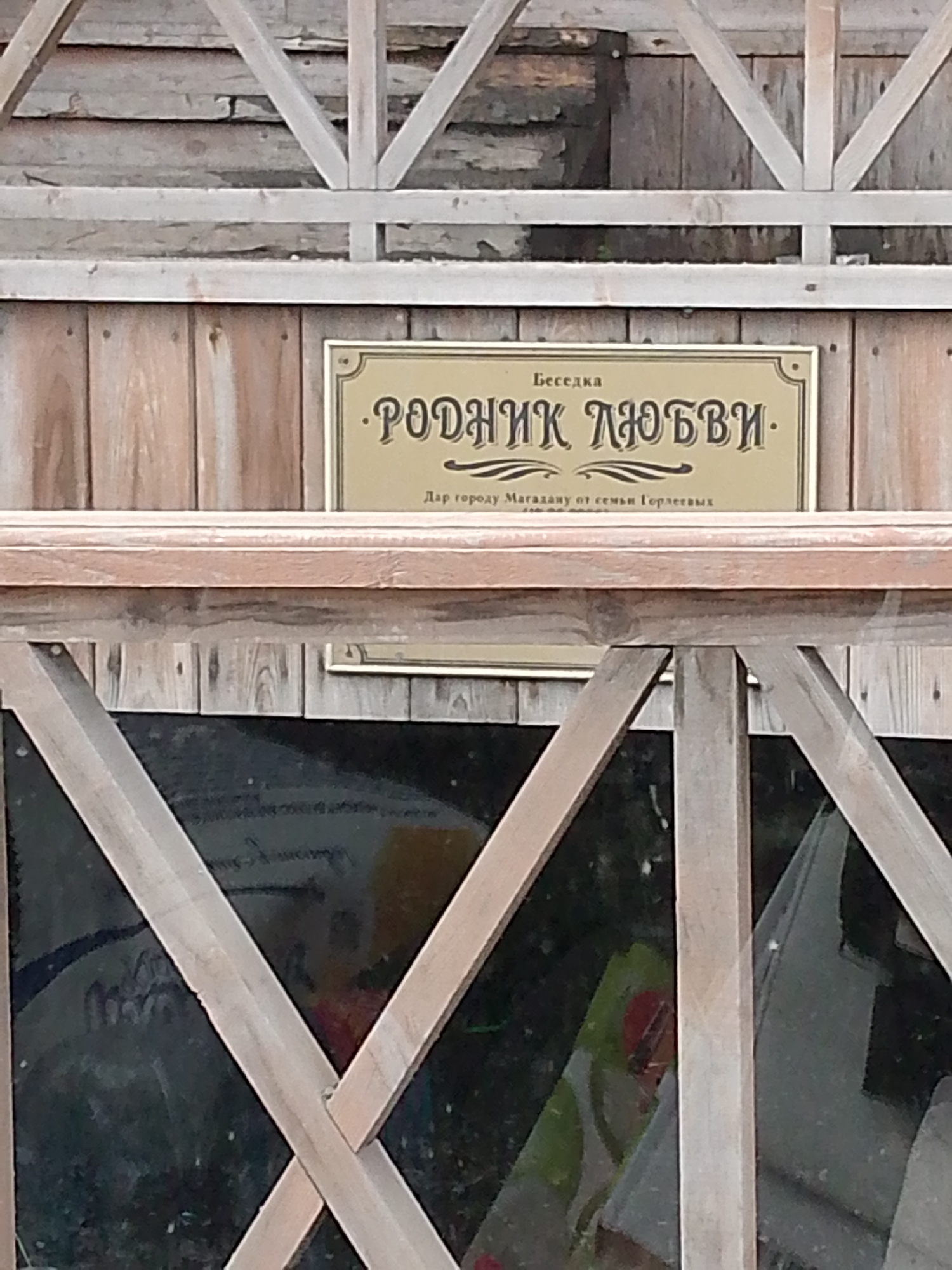 The image shows a sign on a wooden structure. The sign is golden with black lettering and has a decorative border. The text on the sign is in Russian. The top line reads "Беседка" (Gazebo). The main text reads "РОДНИК ЛЮБВИ" (Spring of Love). Below this, in smaller print, it says "Дар городу Магадану от семьи Гордеевых" (Gift to the city of Magadan from the Gordeev family). The background is a wooden wall, and in the lower part of the image, there is a dark reflective surface, possibly glass, showing blurred colorful shapes. The overall scene appears to be outdoors.