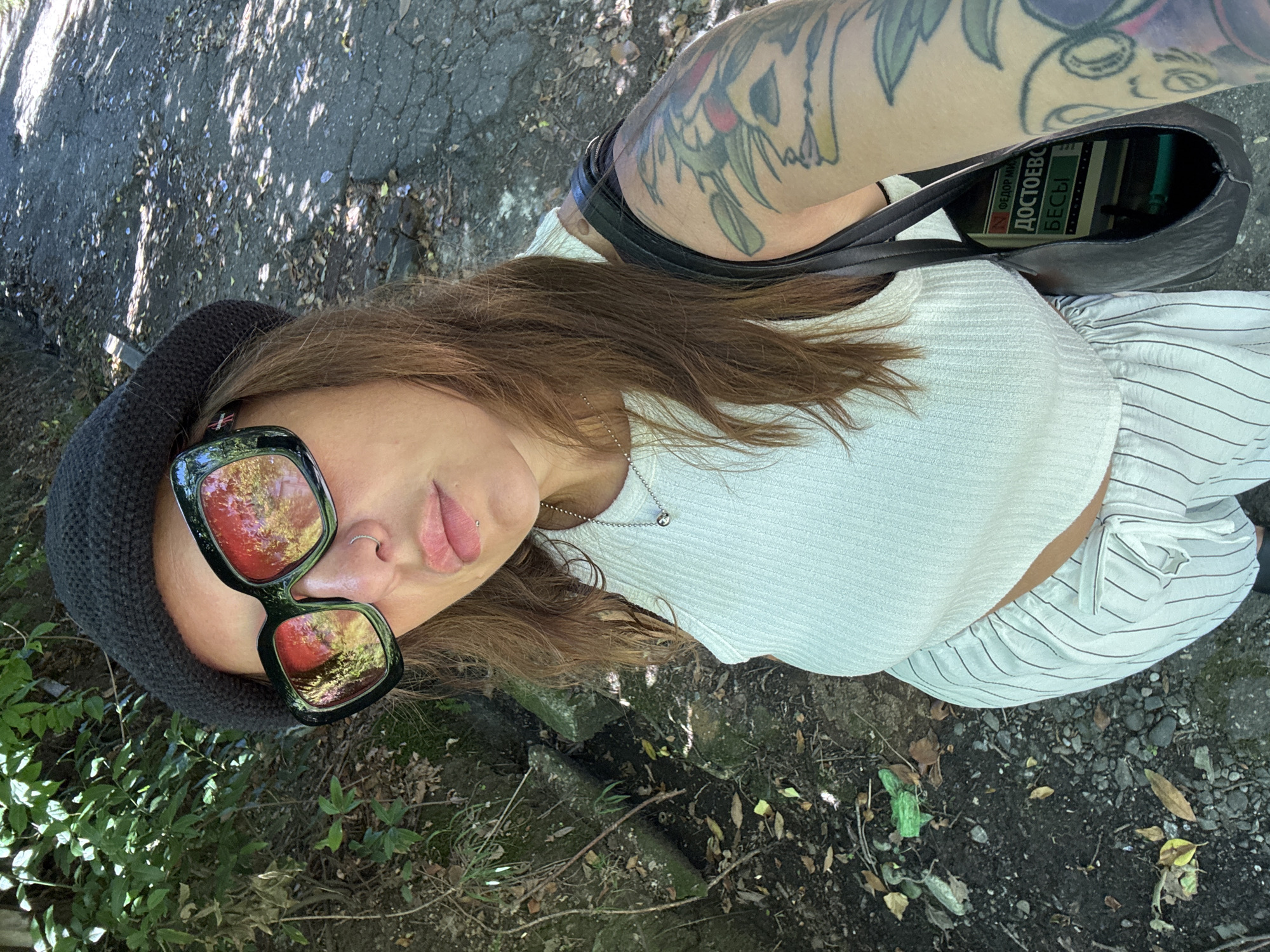 A woman is taking a selfie outdoors in Sochi, Russia. She is wearing a black bucket hat, large square sunglasses with red reflective lenses, a white ribbed crop top, and white pinstripe pants. She has a nose ring and a lip piercing. She is also wearing a silver pendant necklace and has a tattoo on her arm. She is carrying a black bag that contains a book with Russian text that appears to say "ДОСТОЕВСКИ БЕСЫ" (Dostoevsky, Demons). The background shows a path with dappled sunlight, green foliage, and some leaves on the ground. The woman is positioned in the center of the frame, with the path extending to her right and dense greenery to her left.