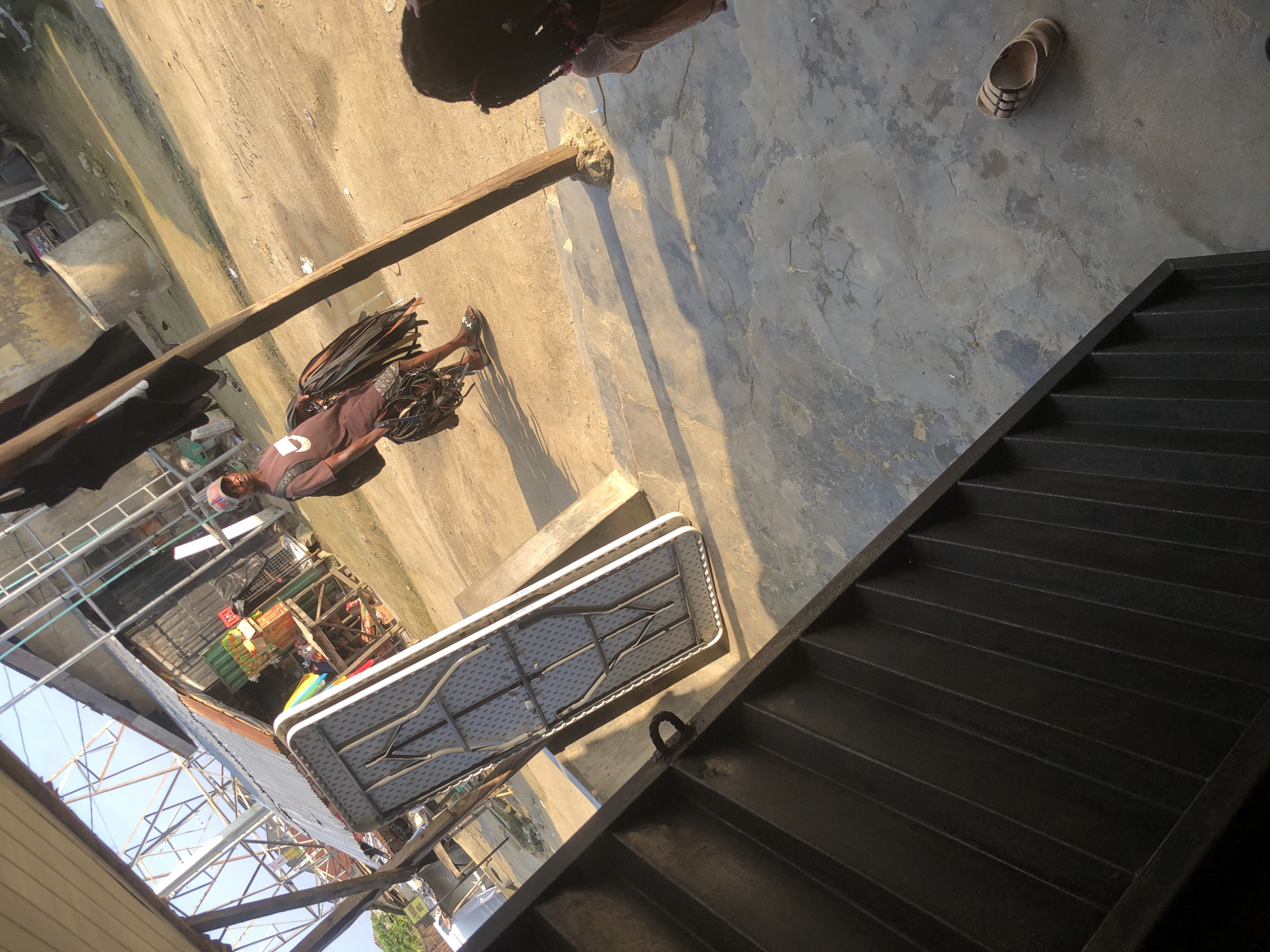 The image depicts a street scene in Ado, Nigeria, under a clear sky. In the foreground, a dark metal staircase with ribbed steps leads diagonally upwards. To the left, a folded table with a patterned surface rests on concrete blocks, casting a long shadow.
A man, wearing a brown t-shirt, shorts, a cap, and flip-flops, walks away from the viewer, carrying a bundle of black cables or ropes. He has a backpack on his shoulders. Behind him, a makeshift stall with corrugated metal roofing and wooden structures is visible, displaying various items, including what appear to be boxes and containers. The ground is a mix of sand and concrete. To the far right, a person's back is visible, with dark hair styled in braids with pink accents. A single flip-flop lies on the concrete ground near the person. The overall environment suggests an outdoor market or a street with commercial activity.