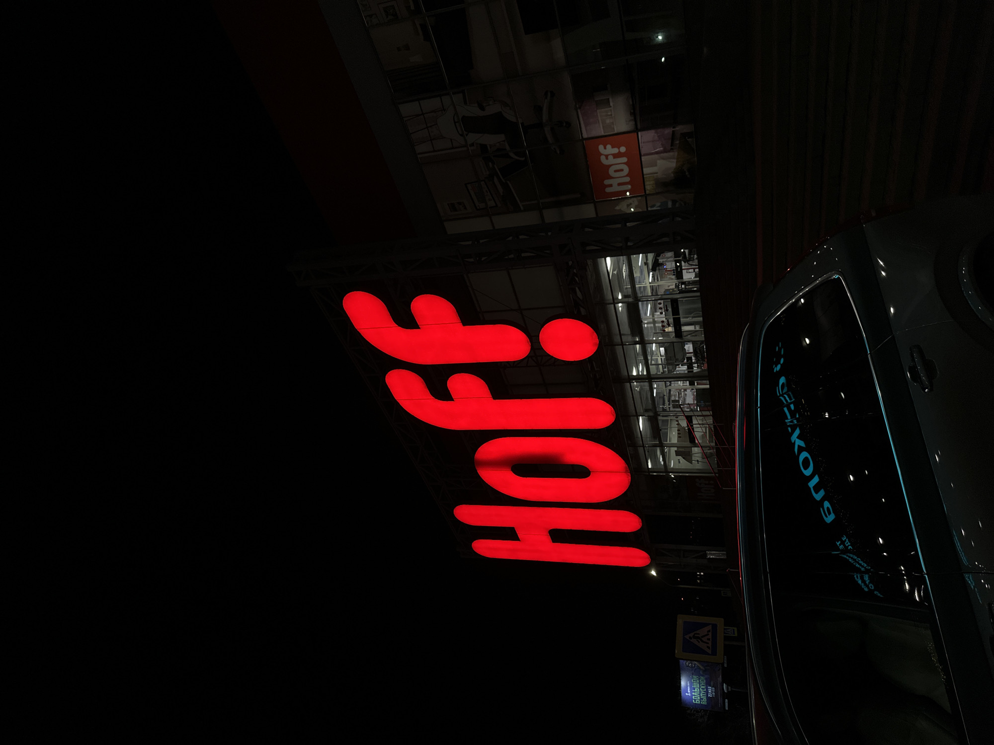 The image depicts the "Hoff" store in Rostov-on-Don, Russia, at night. A large, red, illuminated "Hoff" sign is prominently displayed. The store's facade is visible behind the sign. A portion of a dark-colored vehicle, possibly a car or SUV, is visible on the right side of the frame. There is also a pedestrian crossing sign.