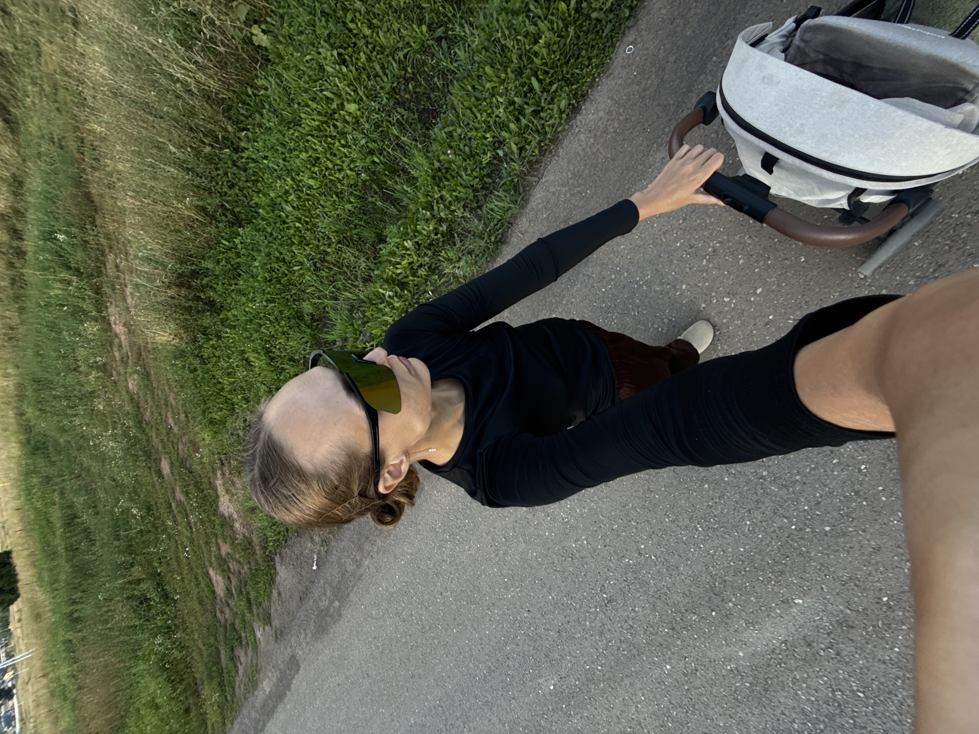 The image shows a first-person perspective of someone walking with a stroller on a paved path next to a grassy area. The person is wearing a black long-sleeved shirt, brown pants, and white shoes. They are holding the stroller's handle. The stroller is light gray with black trim and brown handles. The grassy area appears to be a field or park with tall grass and some wildflowers. The scene suggests it is daytime, possibly late afternoon or early evening, given the soft lighting. The overall mood appears casual and relaxed, indicative of an outdoor stroll. No text is visible in the image. The location is identified as Подшивалово, Russia.