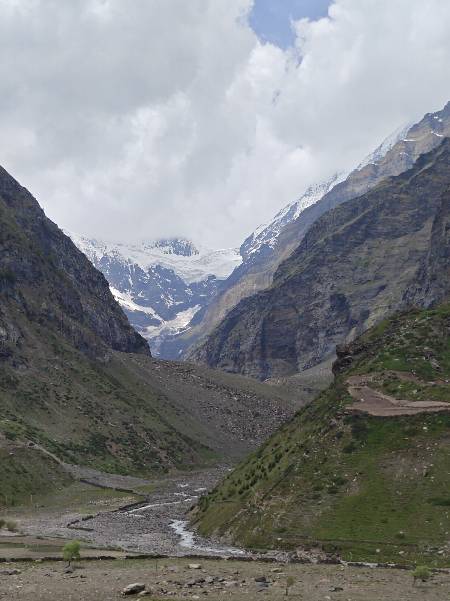 The image depicts a wide mountain valley in Sissu, India. Towering, rugged mountains with steep, rocky slopes dominate the scene, with some areas covered in patches of green vegetation. In the distance, majestic snow-capped peaks rise, their surfaces sculpted by glaciers and snowfields under a cloudy sky. A meandering river flows through the valley floor, its path lined with rocks and stone walls. The valley itself appears to be a natural pathway between the mountains, with a road or trail visible on one of the slopes. The overall environment is wild and remote, characterized by natural geological formations and a cold, alpine climate. There are no people visible in the image.