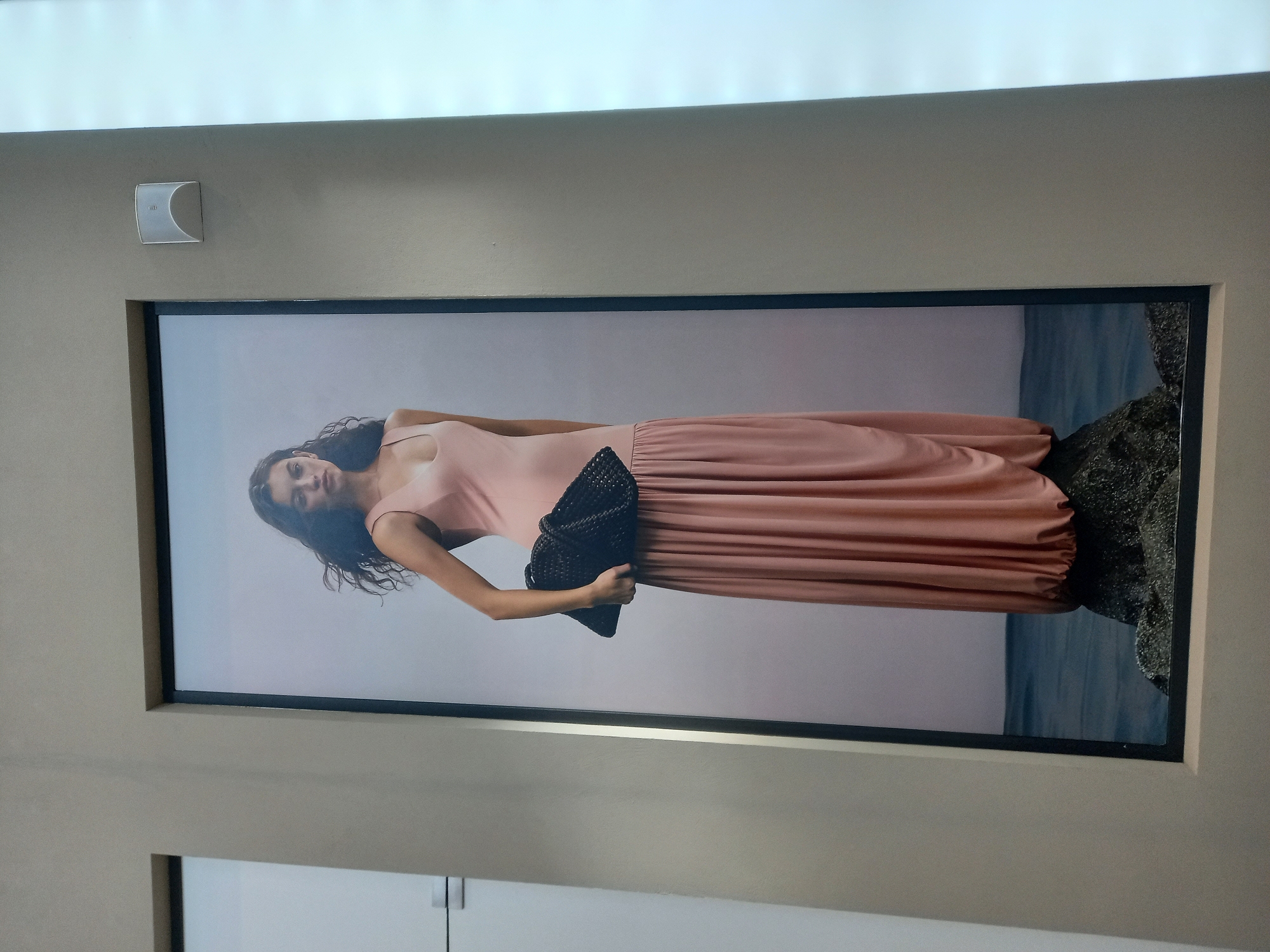 The image shows a framed poster of a young woman wearing a flowing pink dress and holding a black textured clutch bag. She is standing on a rock with water behind her, against a pale pink background. The poster is mounted on a wall in a commercial space, possibly a store, with other adjacent walls visible. To the left of the poster is a white door with a horizontal handle. To the right of the poster, a white illuminated panel is visible. On the wall above and to the right of the poster, a small white motion sensor or alarm device is mounted. The overall lighting suggests an indoor environment. There is no information in the image to suggest a specific location in Yekaterinburg, Russia.