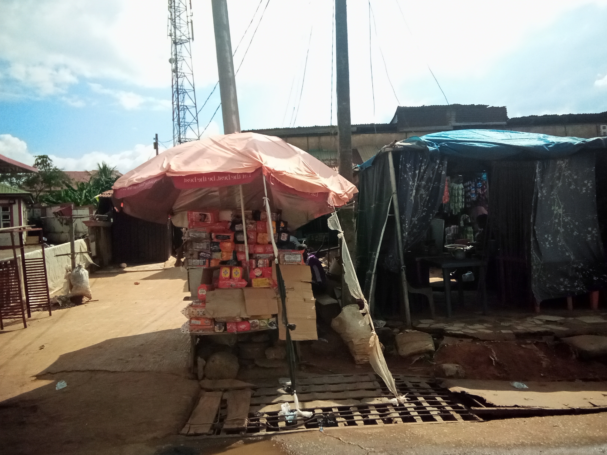 In Nibo, Nigeria, this image depicts an outdoor market scene on a sunny day with a cloudy sky. The foreground features a narrow, unpaved path with a metal grate at the edge. To the left, a small stall is set up under a pink umbrella, displaying various packaged goods, primarily food items like biscuits and snacks, stacked on shelves and boxes. Behind this stall, a white bag and some shelving are visible. To the right, a larger, more permanent stall is covered by a blue tarp, with dark, patterned fabric curtains on the sides. Inside this stall, items are hung and displayed, including what appear to be clothing or textiles, and possibly bottles. A dark table and chairs are partially visible within. In the background, a tall telecommunications tower stands prominently against the sky, with utility wires strung across the scene. Buildings with corrugated metal roofs and wooden structures are also present in the middle ground. The ground is mostly dirt and a lighter-colored paved surface, with some debris scattered around. The overall impression is one of informal commerce in a developing urban or semi-urban area.
