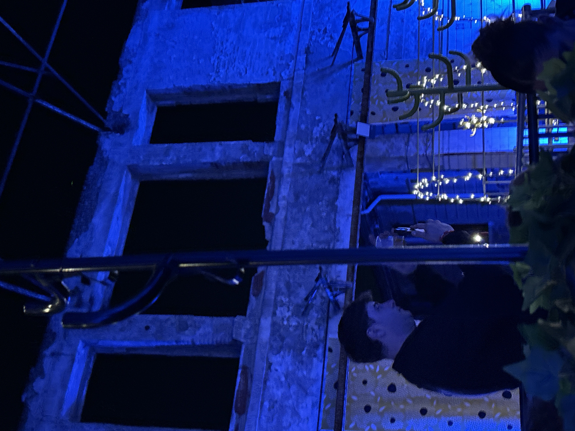 The image shows a man in a dark suit seated at a table in front of a building façade. The building has multiple window openings, some with visible scaffolding. The scene is illuminated with blue light, which suggests that this may be either a nighttime setting or an indoor re-creation. The table is decorated with a spotted pattern and illuminated by string lights. Some greenery is visible in the scene, possibly indicating an outdoor setting or decoration. While not explicitly visible, the location is likely Milan, Italy, given the user context.