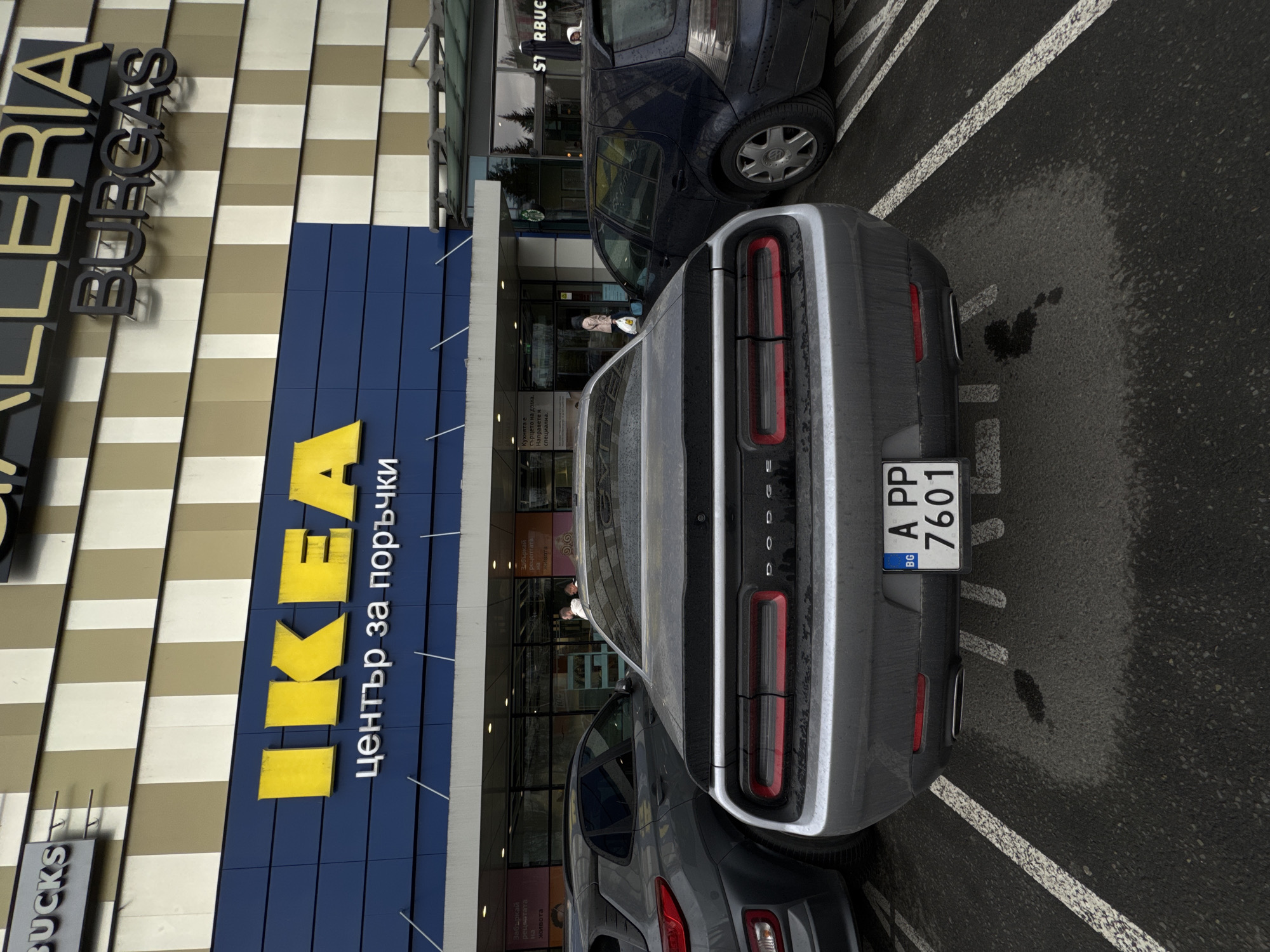 The image shows the rear of a silver Dodge Challenger parked at a parking space outside an IKEA store in Burgas, Bulgaria. The IKEA store is part of a larger Galleria shopping center, as evidenced by the "Galleria Burgas" sign. Next to the IKEA store is a Starbucks outlet. A dark-colored Volkswagen car is parked in front of the Dodge Challenger, while part of another dark-colored SUV is visible behind it. Two people are visible at the entrance of IKEA. The parking lot is wet, with water marks visible on the asphalt. The car's license plate reads "A PP 7601" and has a Bulgarian country code, "BG".