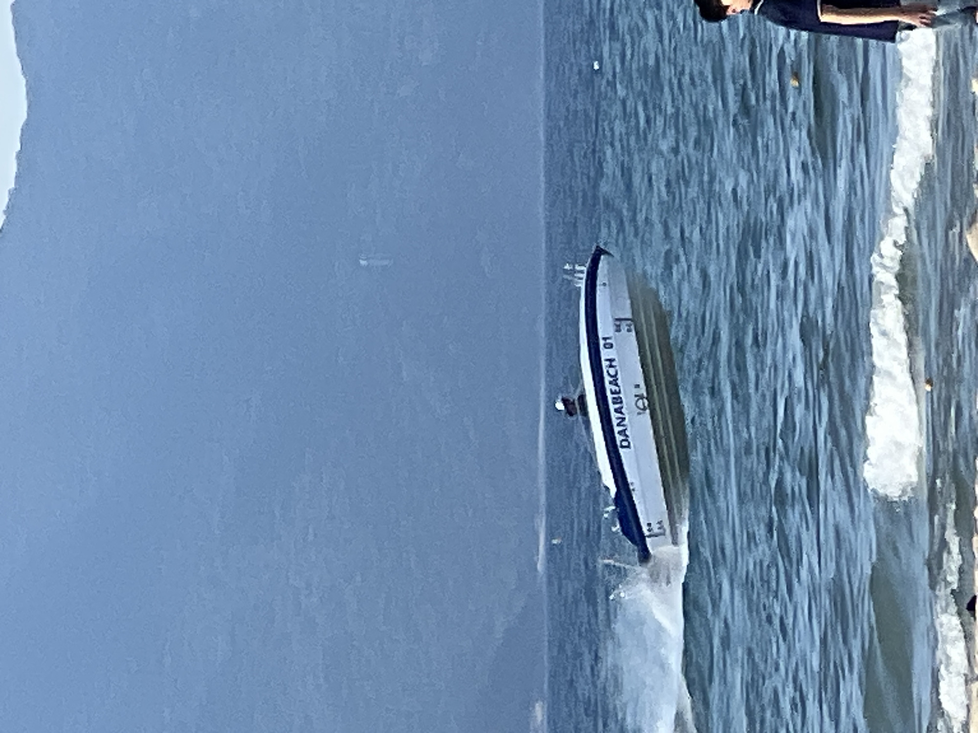In the city of Đà Nẵng, Vietnam, a white and blue speed boat labeled "DANABEACH 01" is seen speeding across choppy blue water, creating a spray of white foam. The boat has a person visible in the driver's seat, wearing a white hat. In the background, a hazy, dark blue mountain range is visible against a light blue sky. In the foreground, on the sandy shore, a person wearing a dark blue t-shirt and light blue jeans stands facing away from the camera, looking out at the water. The waves are breaking on the shore, with some white foam visible.