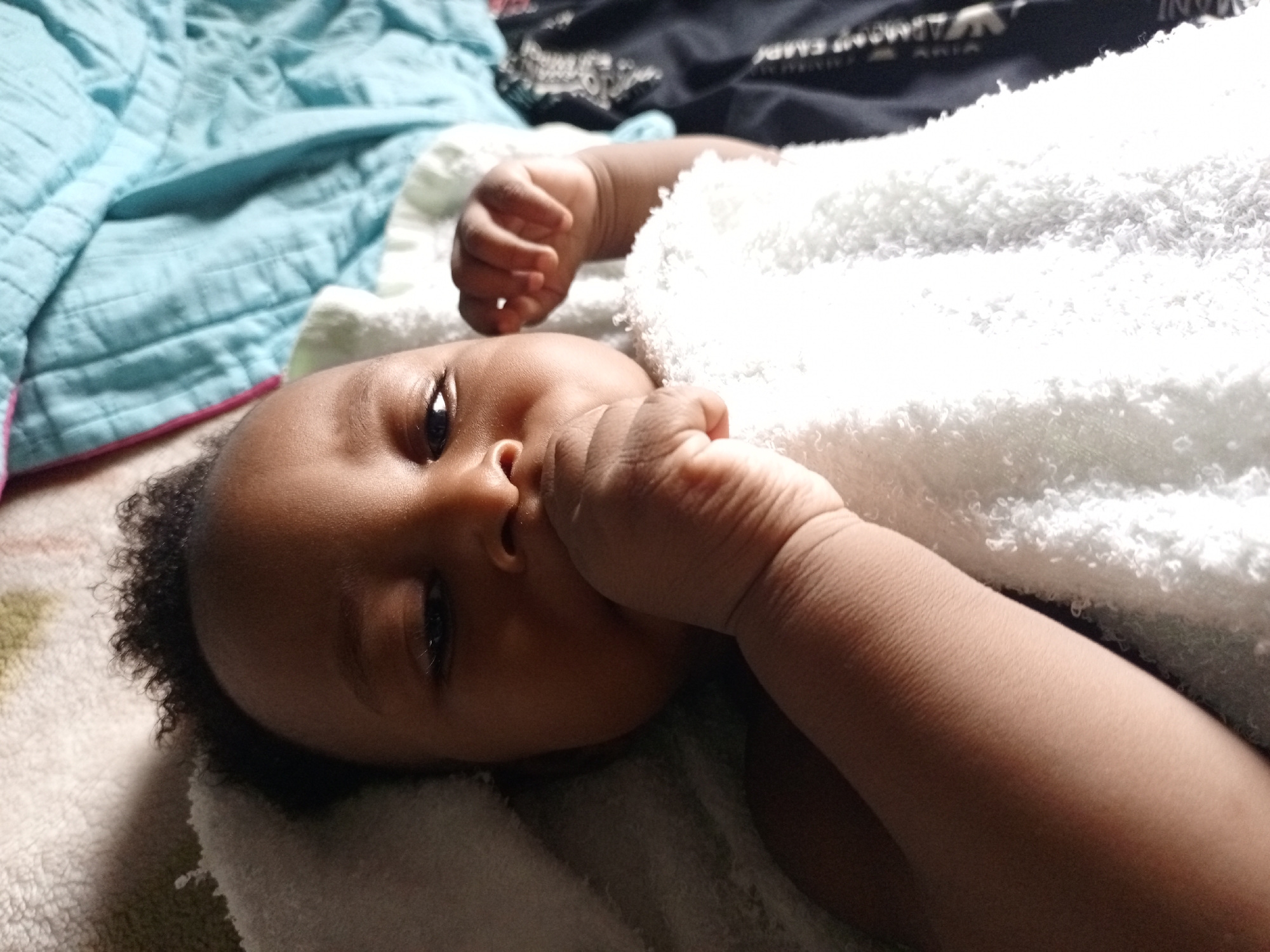 A baby of African descent lies on a patterned blanket, partially wrapped in a white, fluffy towel. The baby's right hand is near their mouth. The background includes a blue blanket and a dark-colored garment with visible text. The location is unspecified, but the request mentioned Keffi, Nigeria.