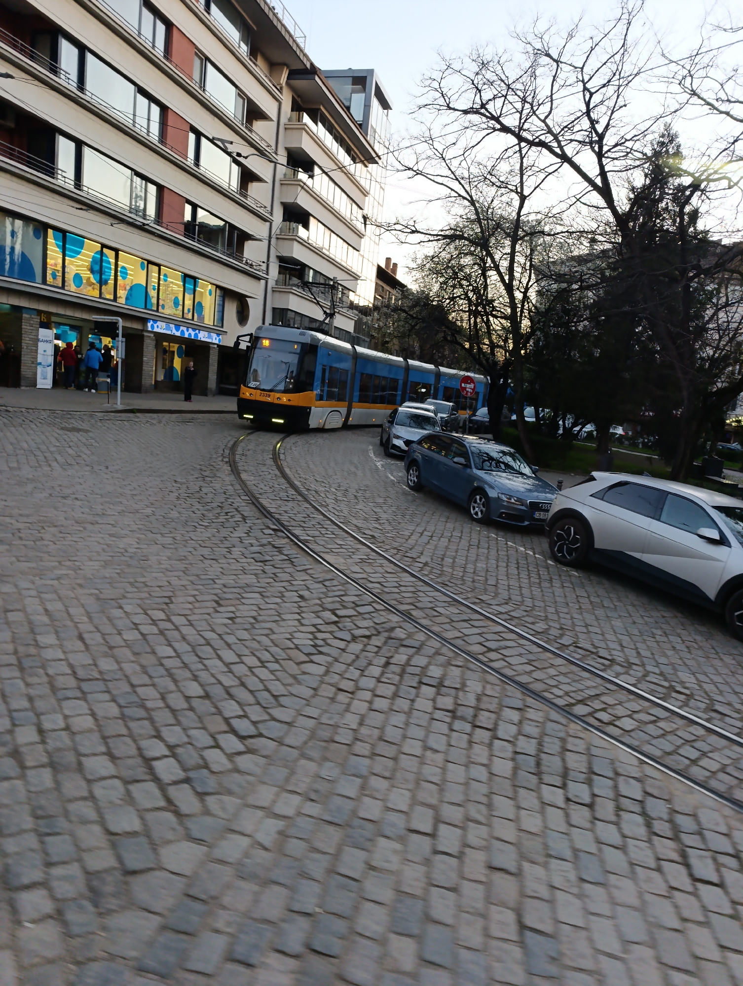 A street scene in Sofia, Bulgaria shows a tram turning a corner on a cobblestone street. In the background, a modern building with glass windows is visible, along with leafless trees. Several cars are parked along the street on the right, including a silver Hyundai Ioniq 5, a gray Audi, and a white van. People can be seen near a storefront, marked with the word "lilly". The tram is light blue and yellow and labeled with the number 18.