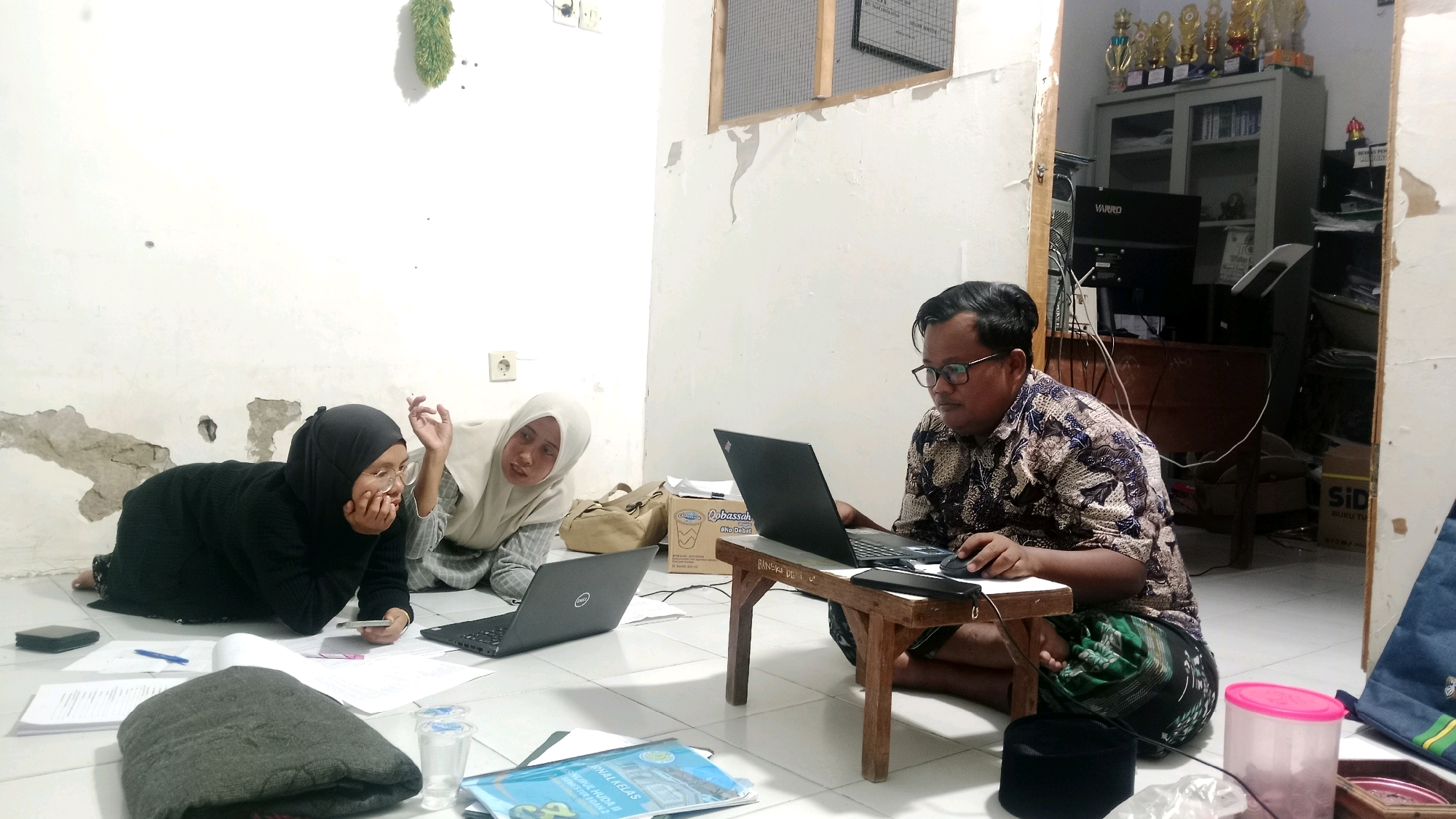 In Dempo Barat, Indonesia, three individuals are engaged in a study session on the floor of a room. A man wearing glasses and a patterned shirt is seated cross-legged in front of a laptop, actively using a mouse. To his left, a woman in a white hijab is also looking at a laptop, her head tilted as if listening or speaking. A third individual, wearing a black hijab and glasses, is lying on her stomach, holding a smartphone and looking at papers spread on the floor.
The environment appears to be a simple room with white walls, some of which show signs of peeling paint. A small wooden table is positioned in front of the man with the laptop. Papers, notebooks, pens, and a folded blanket are scattered on the floor around them. In the background, a cabinet filled with trophies and office supplies, along with computer equipment, suggests an academic or administrative setting. A blue bag and a pink container are also visible in the foreground. The lighting is subdued, indicating an indoor, possibly late-day or evening setting.