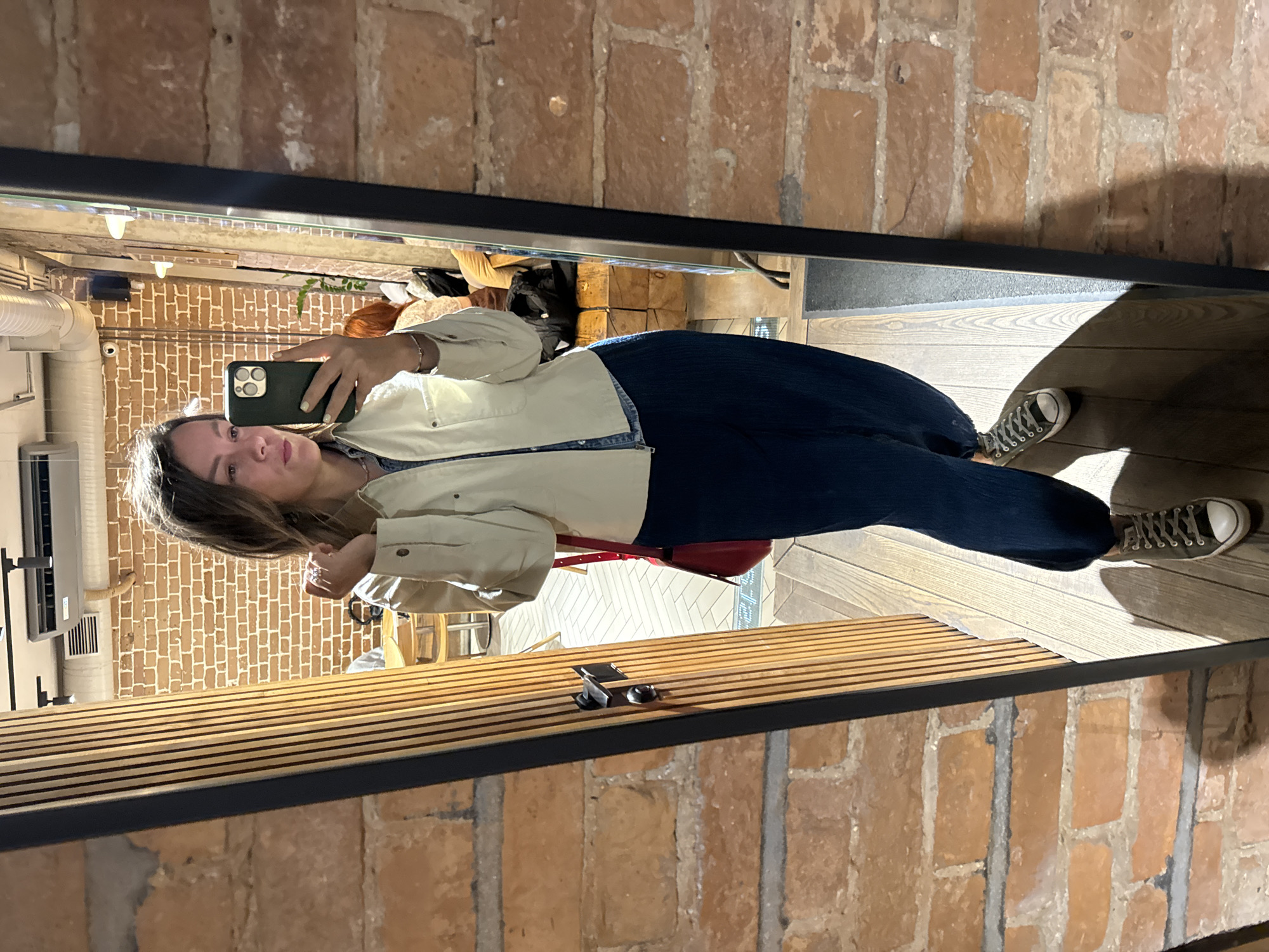A woman stands in a room with exposed brick walls, taking a mirror selfie. She is wearing a light-colored jacket over a denim shirt, dark blue wide-leg pants, and dark sneakers. A red purse is slung over her shoulder. The room appears to be a cafe or restaurant, with wooden paneling, a door, and visible patrons in the background. The image is undated and unlocated.