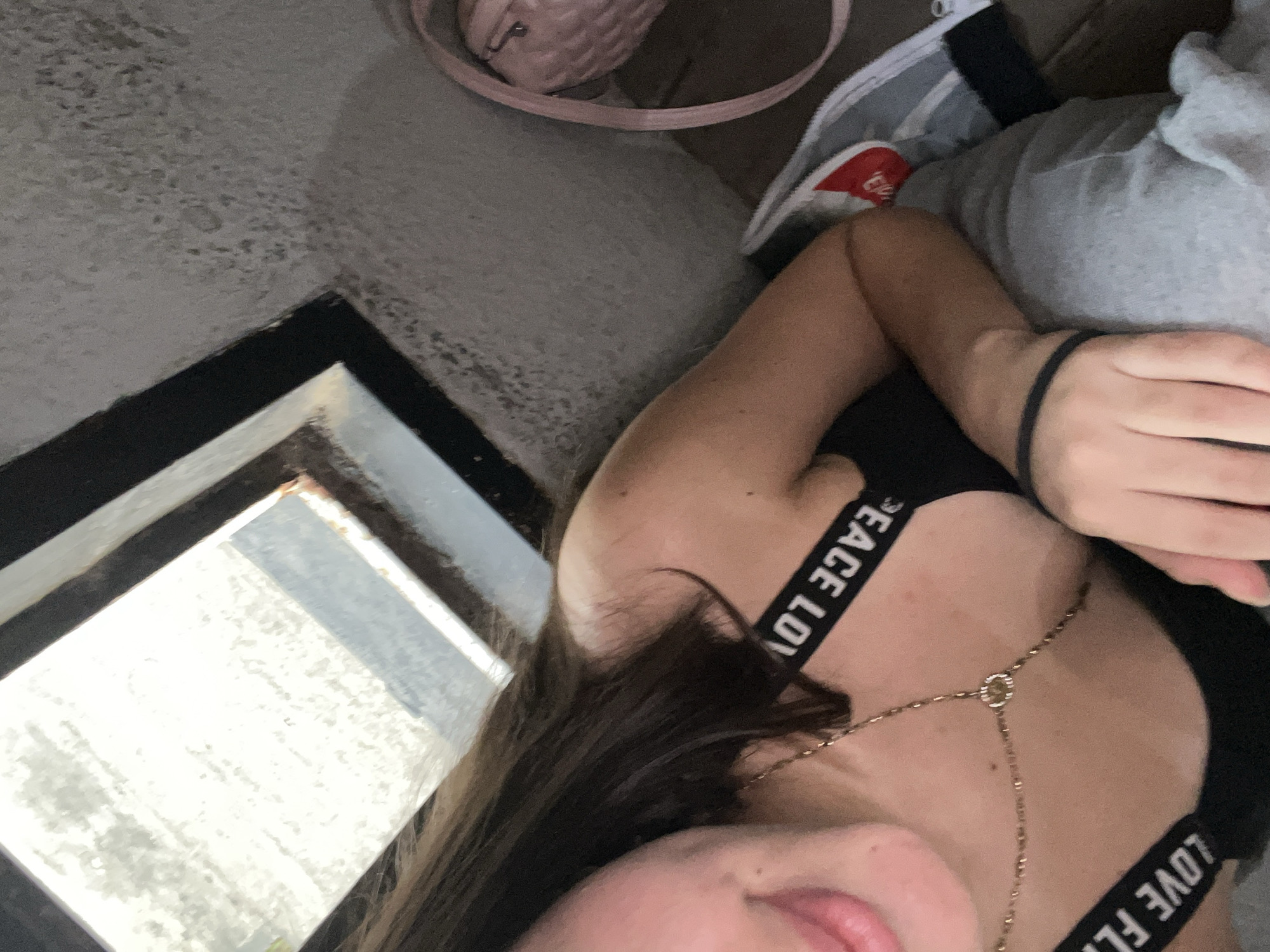 The media file captures a close-up, rotated view of a person, likely female, in an indoor setting in Juan Griego, Venezuela. The subject's upper torso, right arm, and a portion of her face (chin and mouth) are visible. She is wearing a black top with wide shoulder straps. A delicate, gold-colored body chain or necklace with a central circular embellishment drapes across her chest. A black elastic band is on her right wrist.
In the background, a light pink quilted handbag with a strap is partially visible towards the upper right. Adjacent to the person, there's a gray fabric, possibly a blanket or clothing. A bright, rectangular window frame is prominent in the lower left, showing an intensely bright, overexposed outdoor view, indicating it is daytime. The surface next to the window and behind the subject appears to be a light grey, textured wall or floor.
The person is in a relaxed, possibly reclining or lounging pose, with her lips slightly parted. The words "PEACE LOV" are visible in white lettering on the upper black strap, and "LOVE FL" is visible on the lower black strap of her top. The image is captured from an overhead or high-angle perspective, rotated approximately 45-60 degrees clockwise.