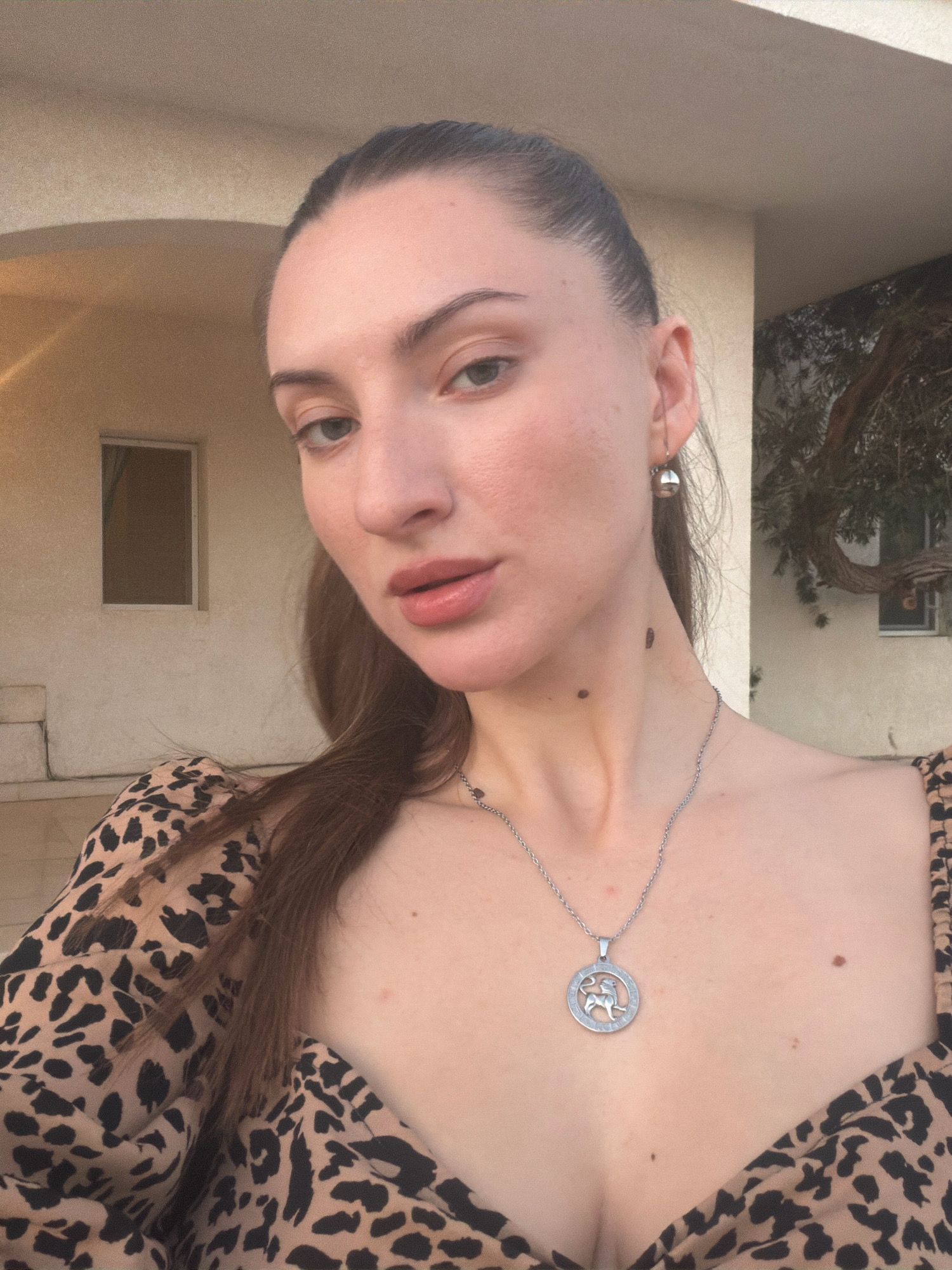 The image is a selfie of a woman in Sharm El Sheikh, Egypt, taken outdoors. The woman is positioned in the center of the frame, facing the camera with her head slightly tilted. She has her hair pulled back into a ponytail and is wearing a leopard-print top. She is also wearing a silver necklace with a circular pendant featuring a lion symbol, and a small silver earring. Her skin appears smooth and fair, with a few moles visible on her neck. The background shows a building with a textured beige facade, a small rectangular window, and some greenery from a tree. The lighting suggests it is either late afternoon or early evening.