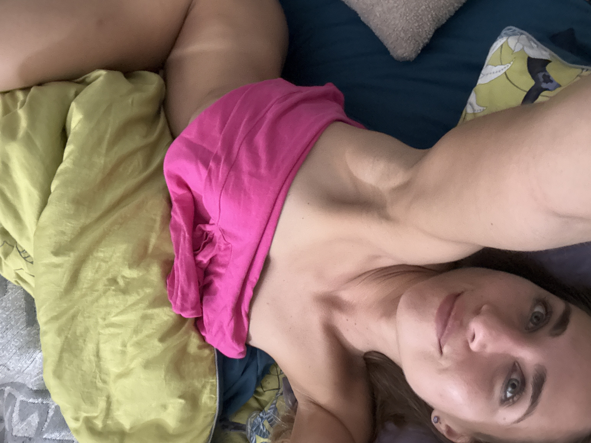 A woman is lying on a bed, looking up at the camera. She is wearing a pink garment. The bed is covered with a green pillow and other bedding. The scene is indoors.